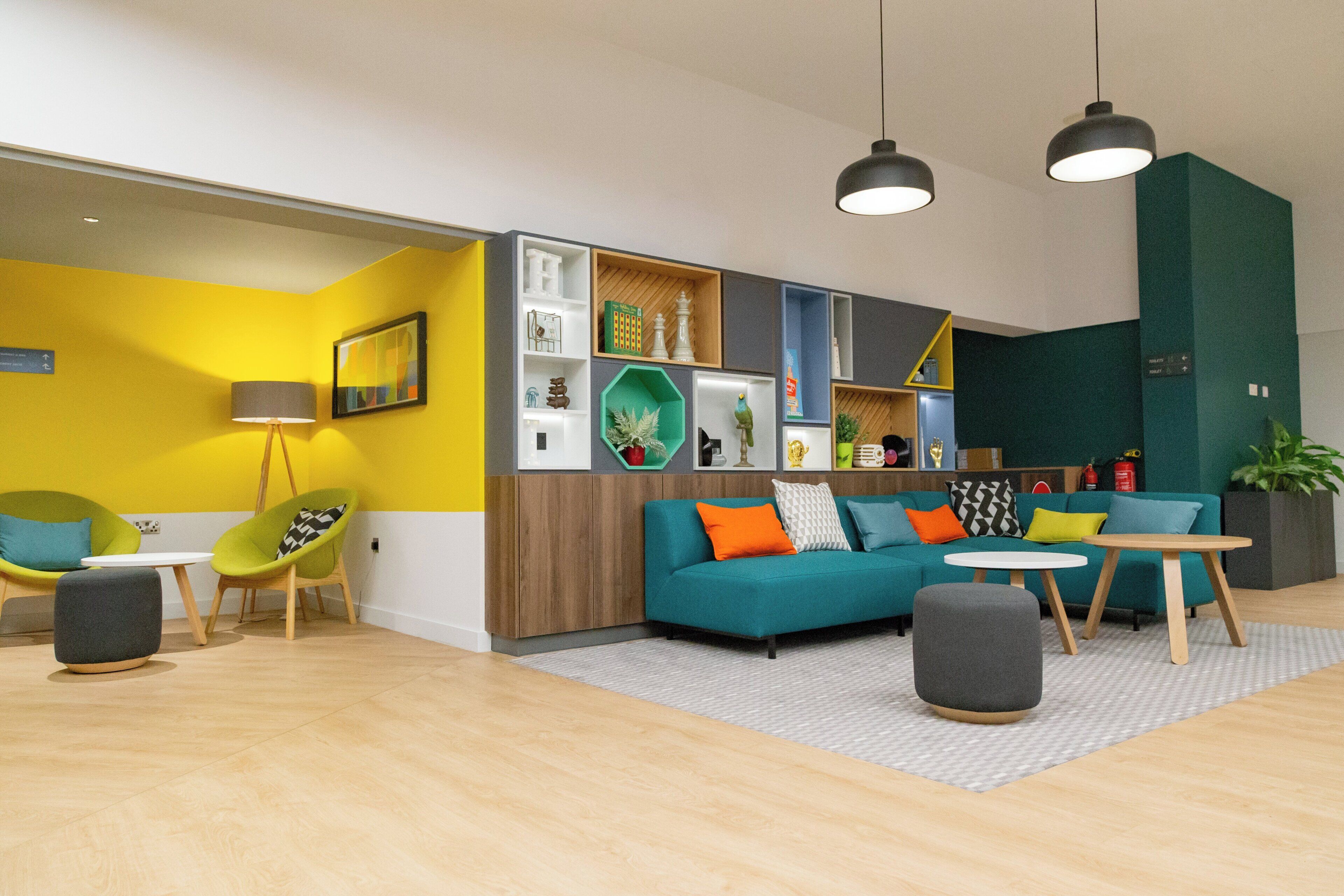Un salon moderne avec un canapé turquoise, des murs jaunes et un plancher en bois. On y trouve deux lampes suspendues, une table basse et une petite table d’appoint.