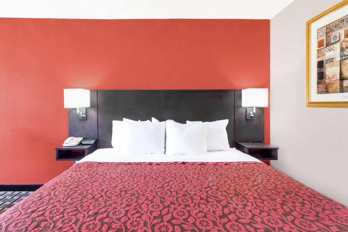 A hotel room with a red accent wall, a large bed with white bedding, two bedside tables with lamps, and a framed artwork on the wall.