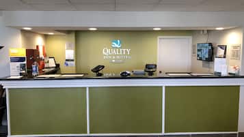 Un comptoir d’accueil d’hôtel aux couleurs vert et blanc, arborant le logo de « Quality Inn & Suites », et offrant divers équipements, tels qu’un distributeur d’eau, un téléphone et un écran d’ordinateur.