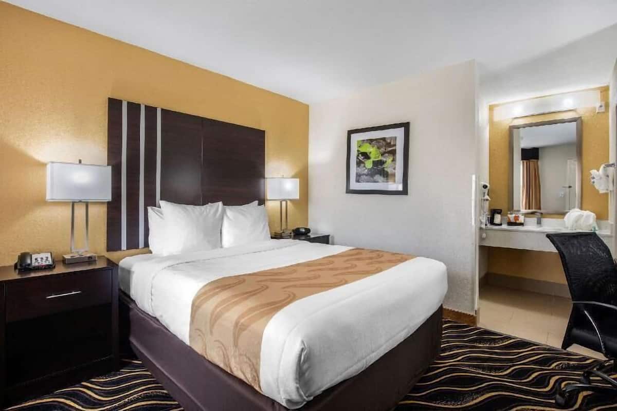 A hotel room with a bed, bedside tables, a desk, a chair, a mirror, and a framed picture on the wall.