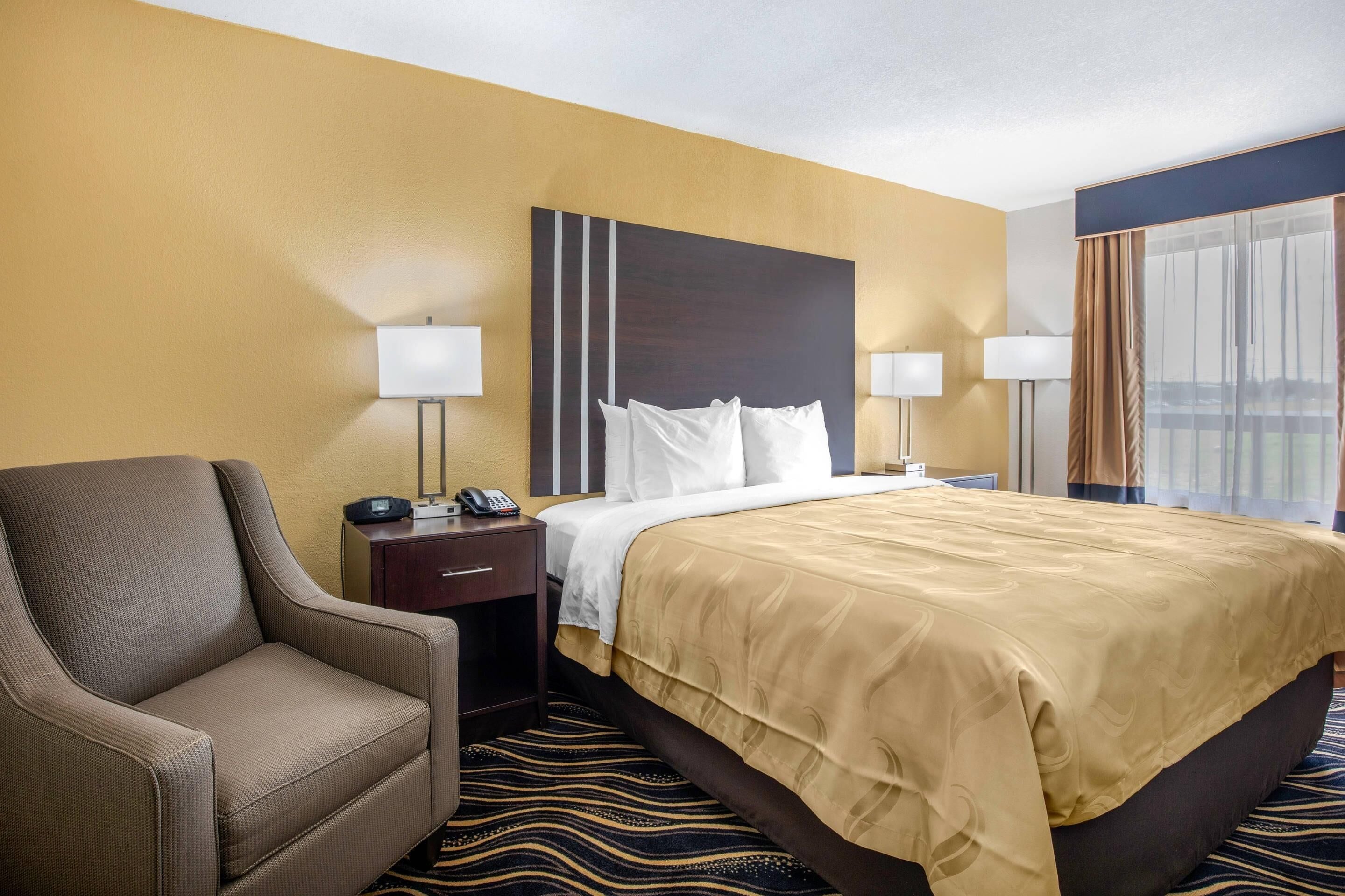 Une chambre d’hôtel avec un lit, une chaise, une table de chevet, une lampe et une fenêtre avec des rideaux.