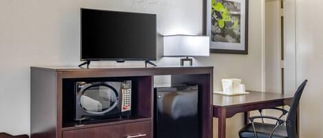 A hotel room with a bed, a desk with a television, a microwave, a refrigerator, a chair, and a framed picture on the wall.