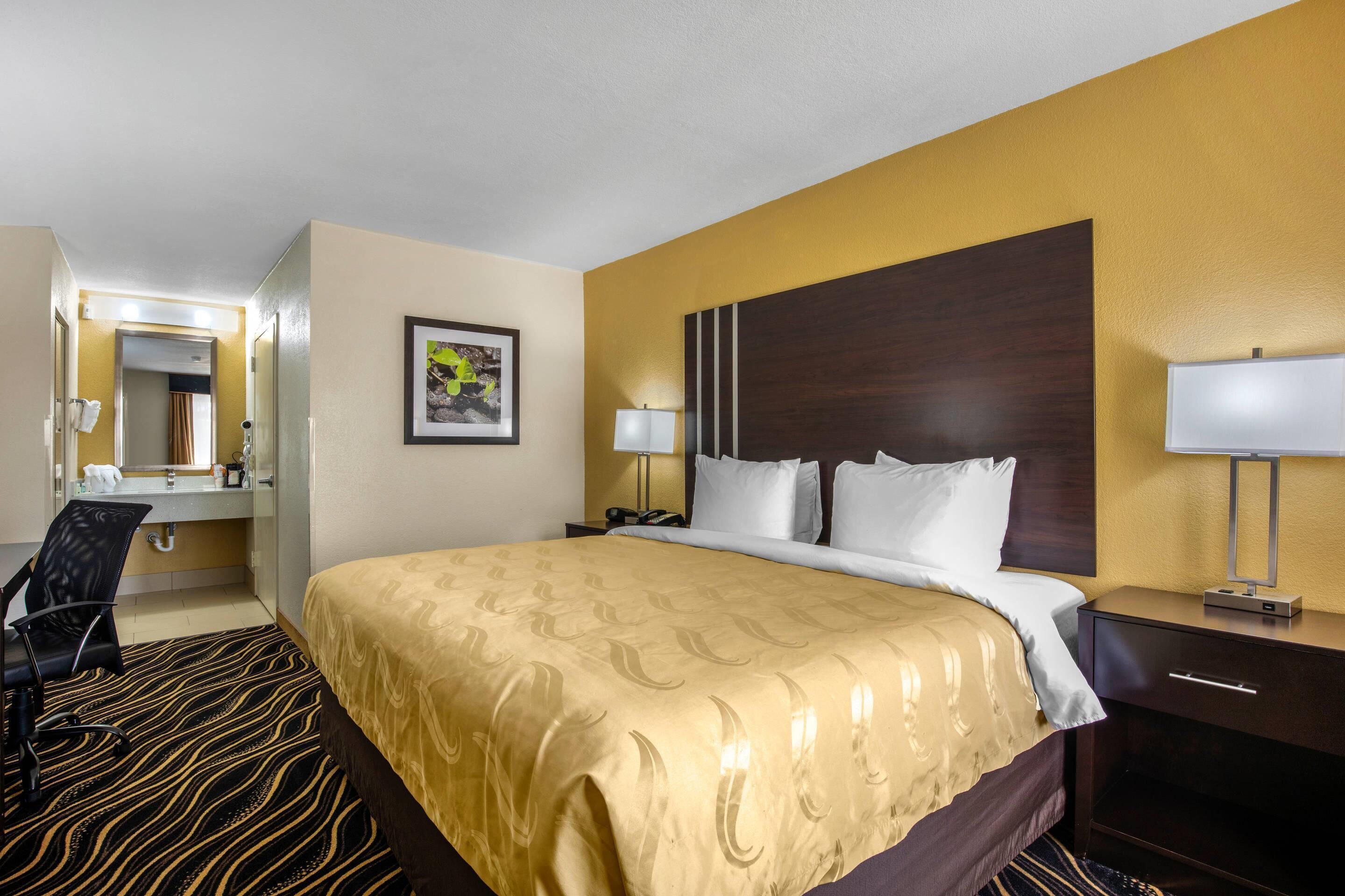 A hotel room with a large bed, a desk with a chair, a mirror, and a bedside table with a lamp.