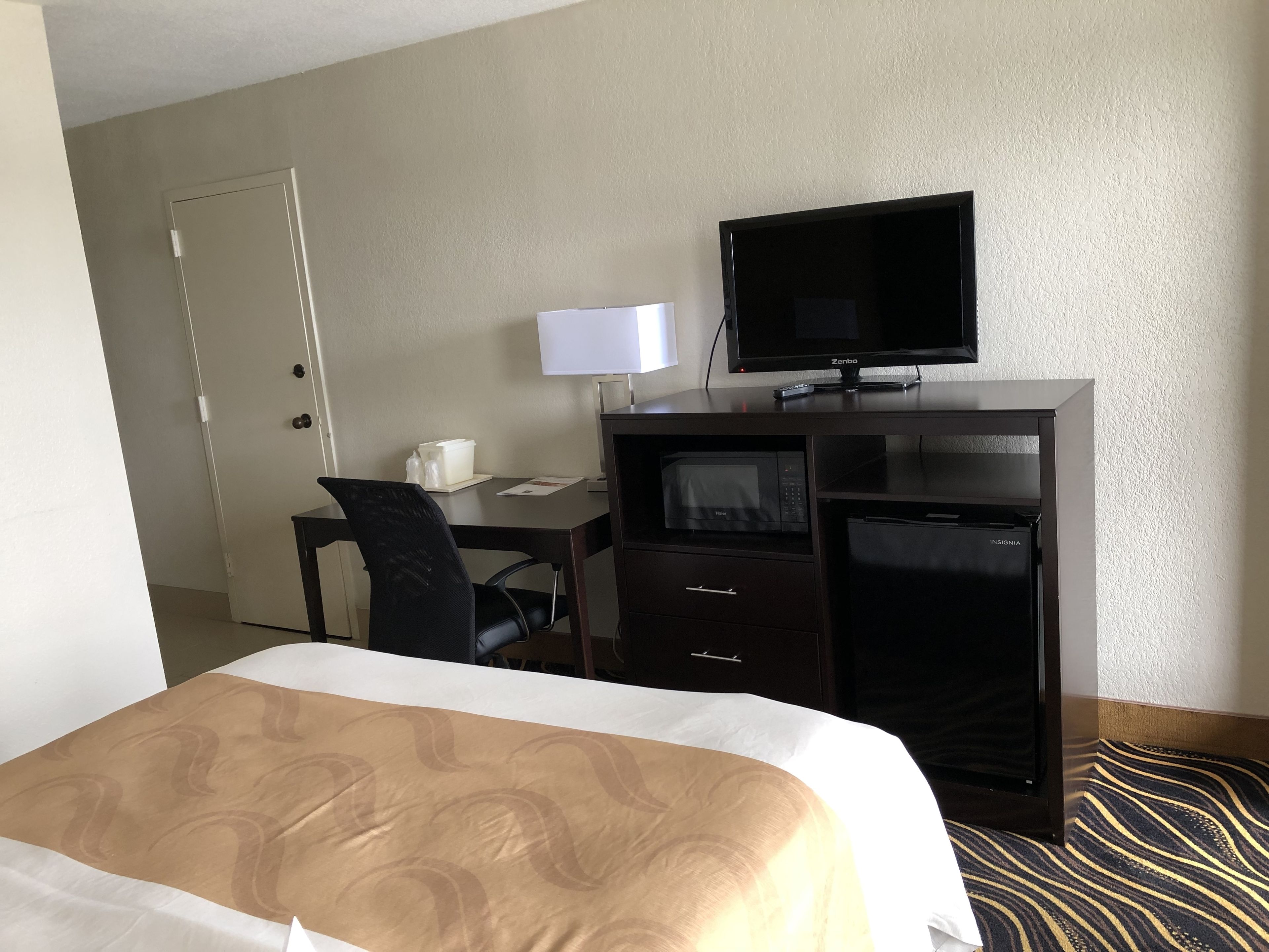 Habitación de hotel con cama, escritorio, computadora, televisor, microondas y minibar.