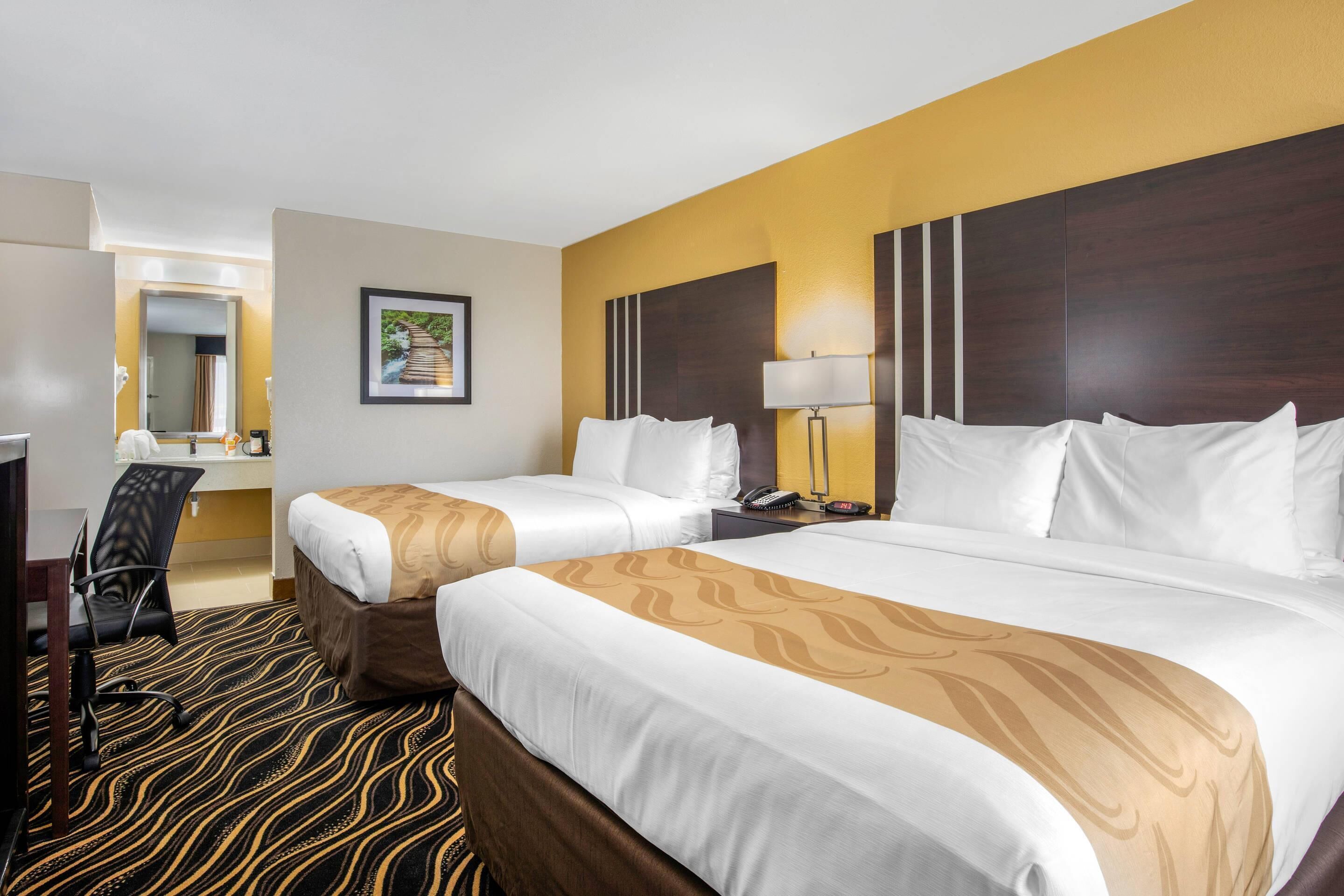 A hotel room with two beds, a desk, a chair, a lamp, and a painting on the wall.