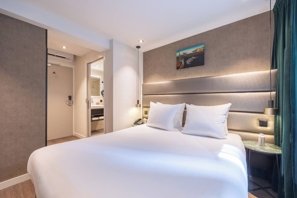 A modern hotel room with a large bed, bedside tables, a painting on the wall, and an air conditioning unit.