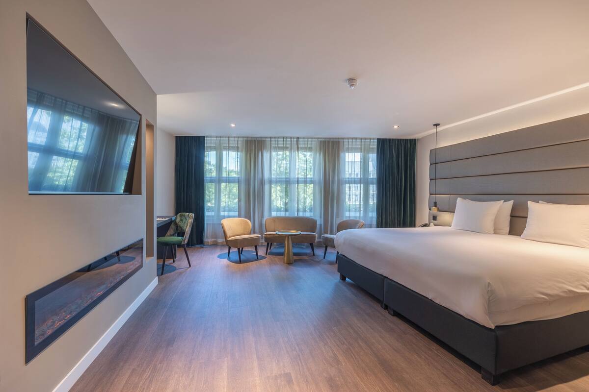 A modern hotel room with a large bed, a seating area with chairs, a small table, and a large window with curtains.