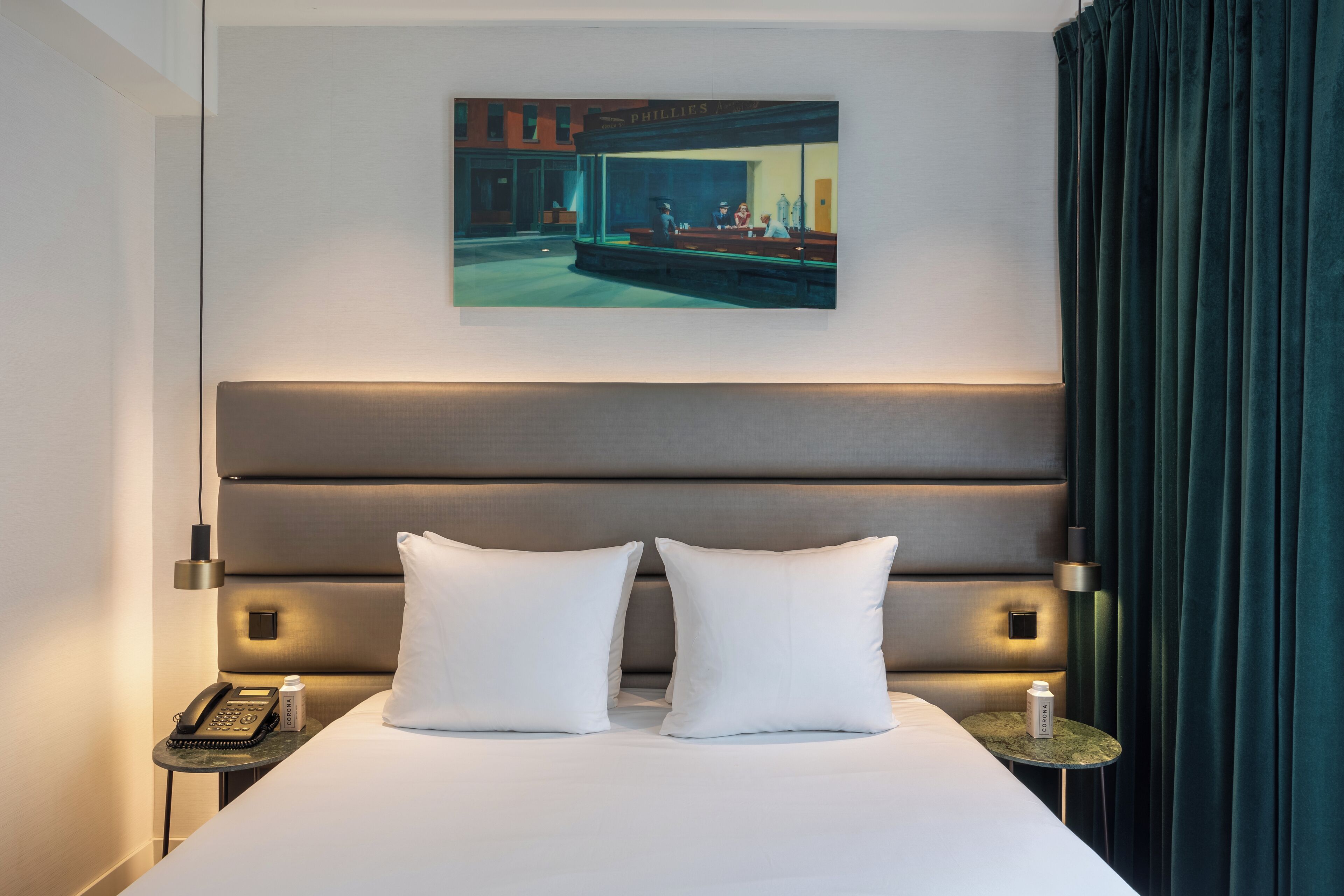 A hotel room with a large bed, two white pillows, a headboard, a bedside table with a phone, a bottle, and a painting of a diner scene on the wall.