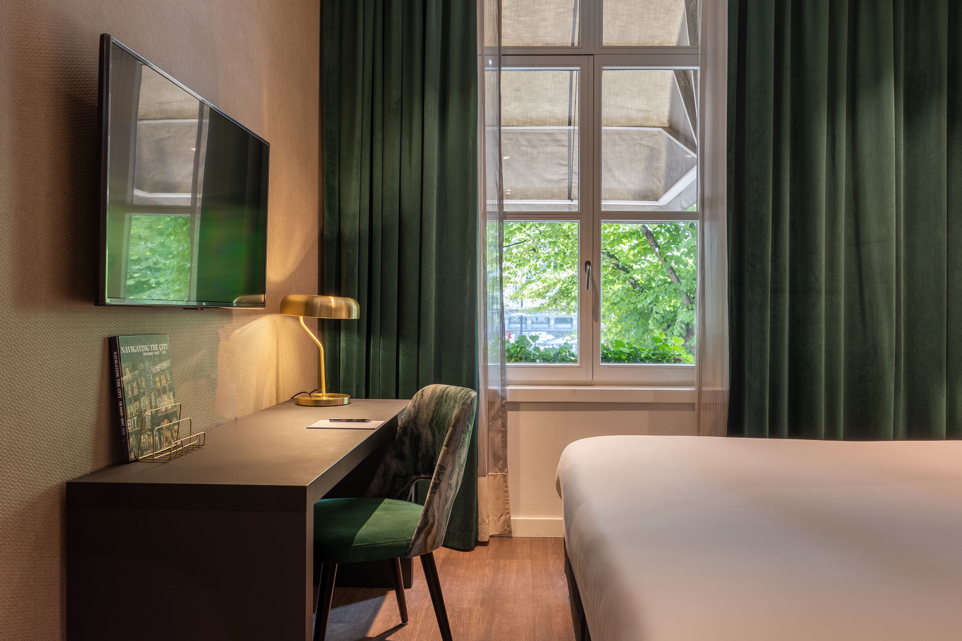A hotel room with a bed, a desk with a chair, a TV mounted on the wall, and a window with curtains.