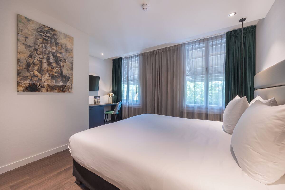 A modern hotel room with a large bed, a desk, and a painting on the wall.