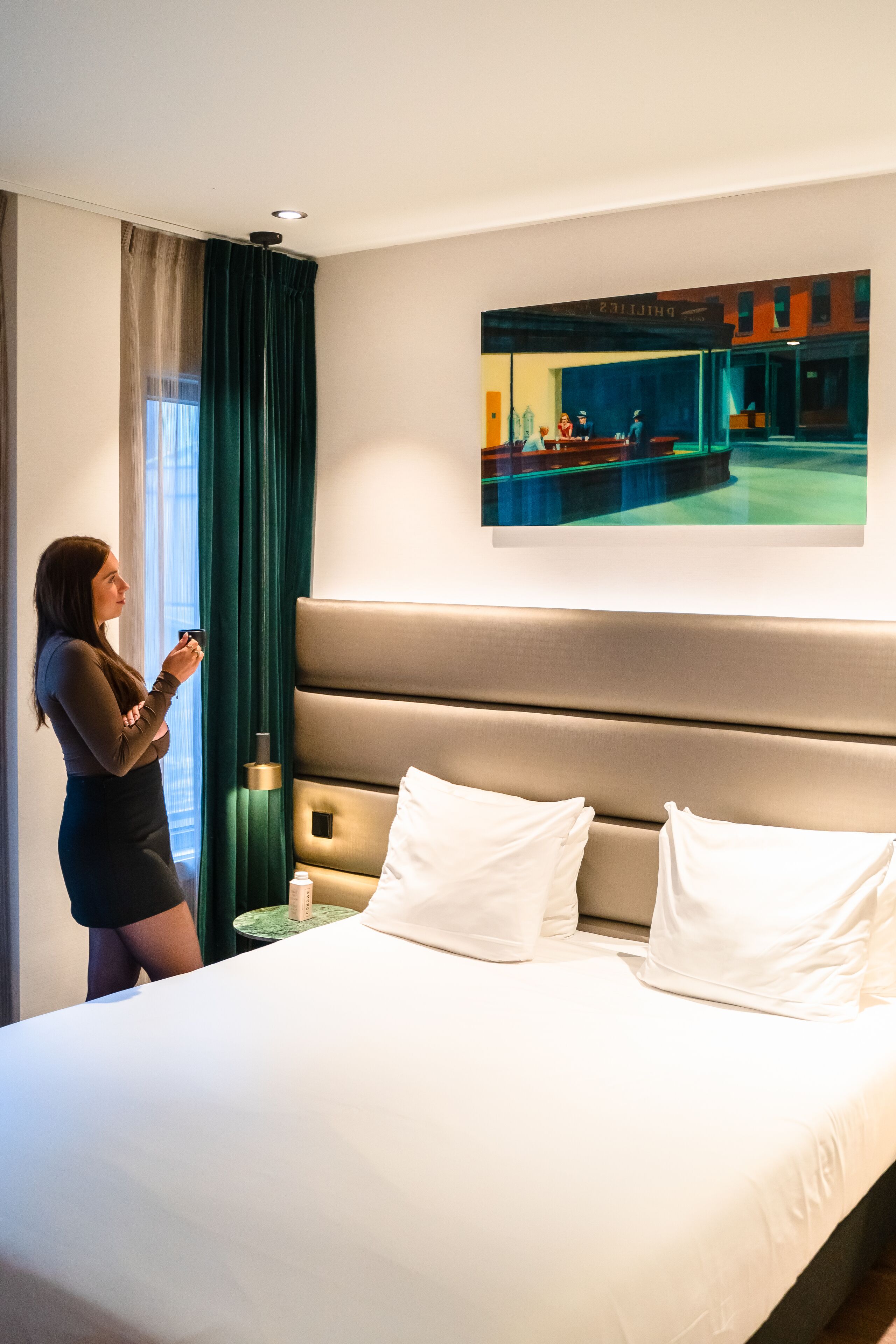 A hotel room with a bed, a nightstand, a painting on the wall, and a window with curtains.