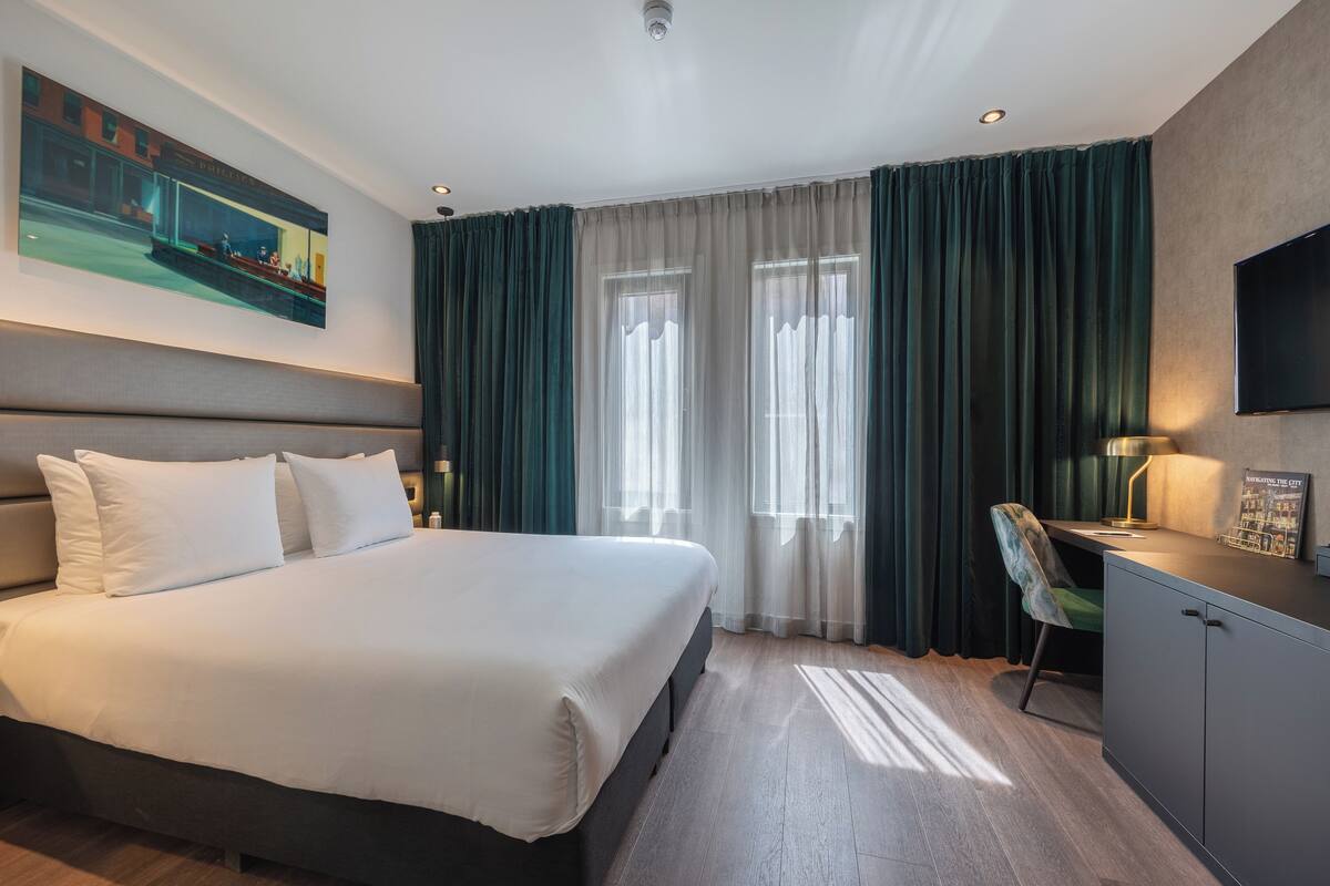 A hotel room with a large bed, a desk, a chair, and a television on the wall.