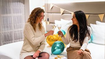 A bedroom with a bed, pillows, and a bedside table with a glass of champagne and balloons.