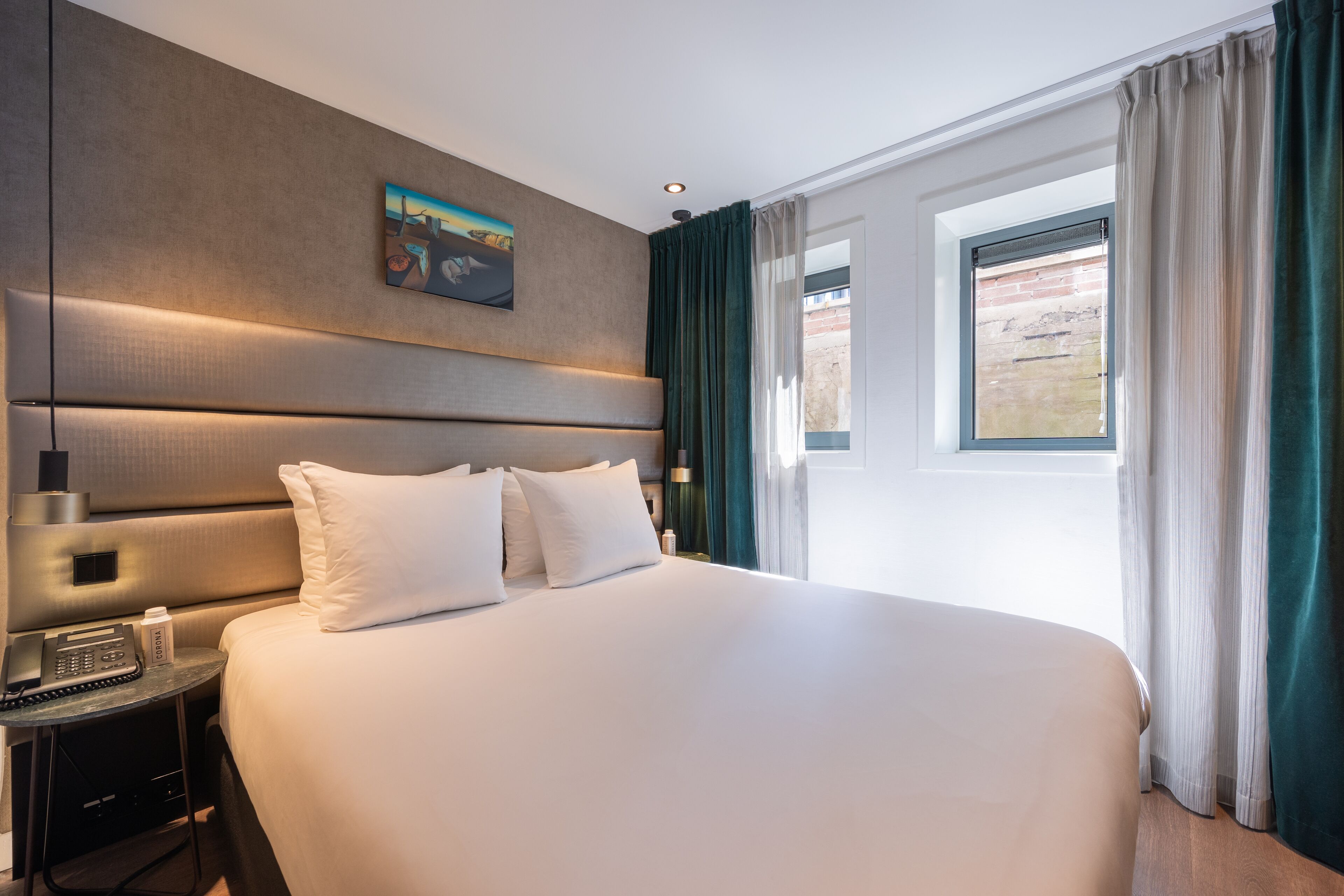 Une chambre d’hôtel avec un grand lit, une table de chevet avec un téléphone, un tableau au mur et une fenêtre avec des rideaux.