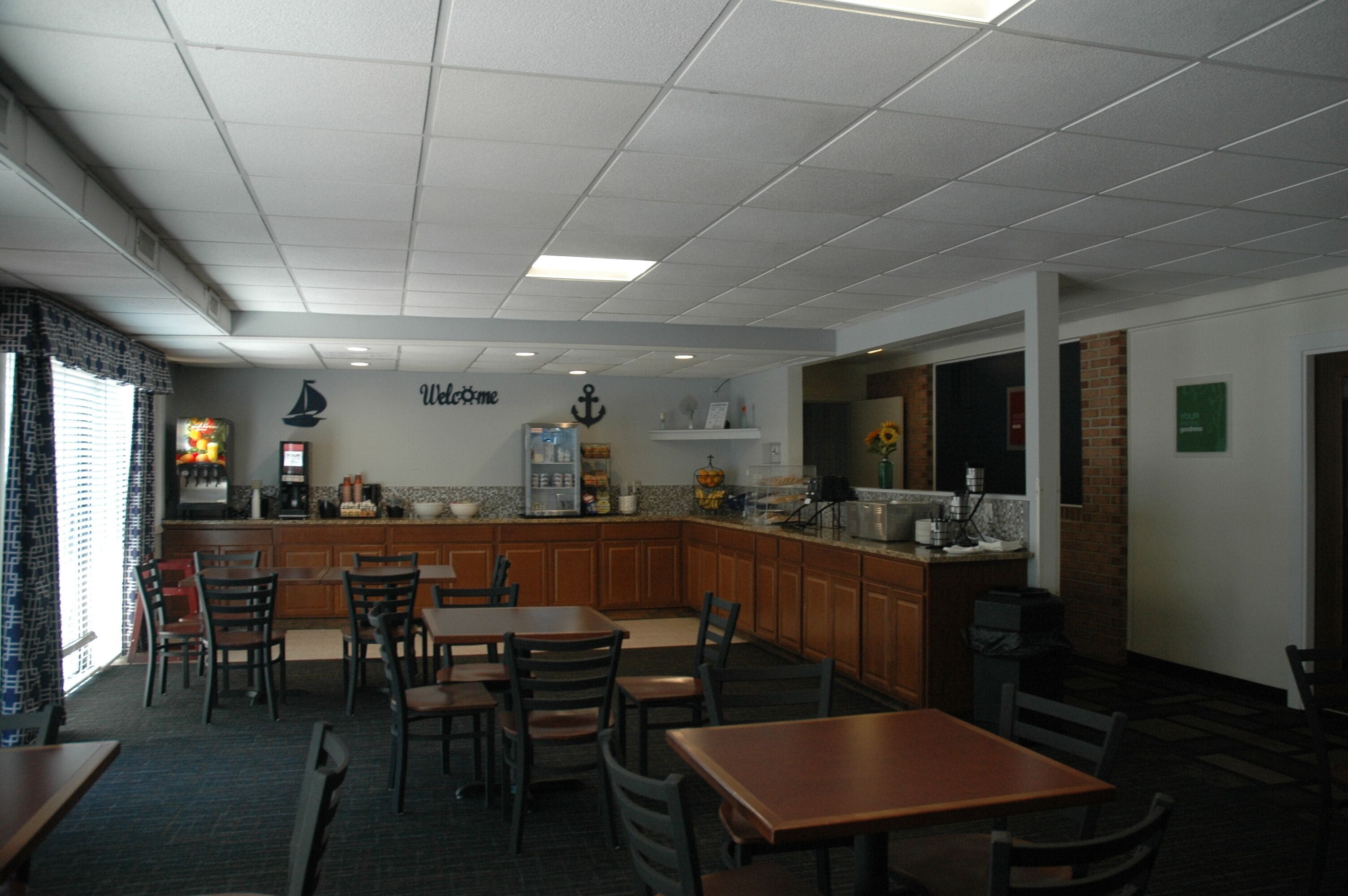 A restaurant with a kitchen area, wooden tables and chairs, and a 'Welcome' sign on the wall.