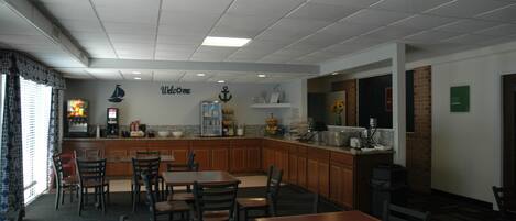 A restaurant with a kitchen area, wooden tables and chairs, and a 'Welcome' sign on the wall.