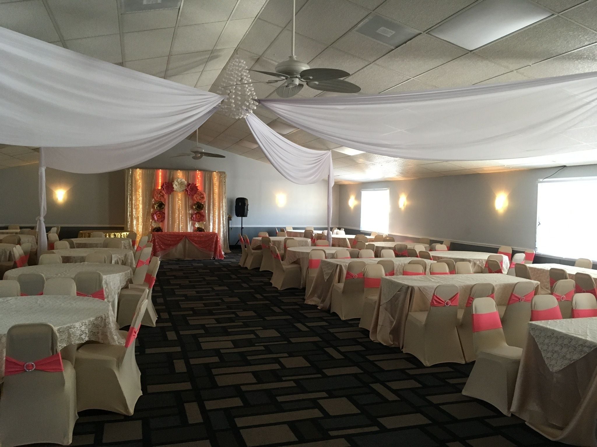 A banquet hall with round tables, chairs with pink sashes, a stage with floral decorations, and a ceiling fan.
