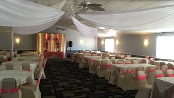 A banquet hall with round tables, chairs with pink sashes, a stage with floral decorations, and a ceiling fan.