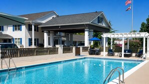 A rectangular swimming pool with a white edge, a covered patio area with outdoor furniture, and a flagpole with two flags. - St Michaels Inn (St. Michaels)