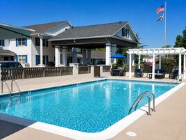 A rectangular swimming pool with a white edge, a covered patio area with outdoor furniture, and a flagpole with two flags.