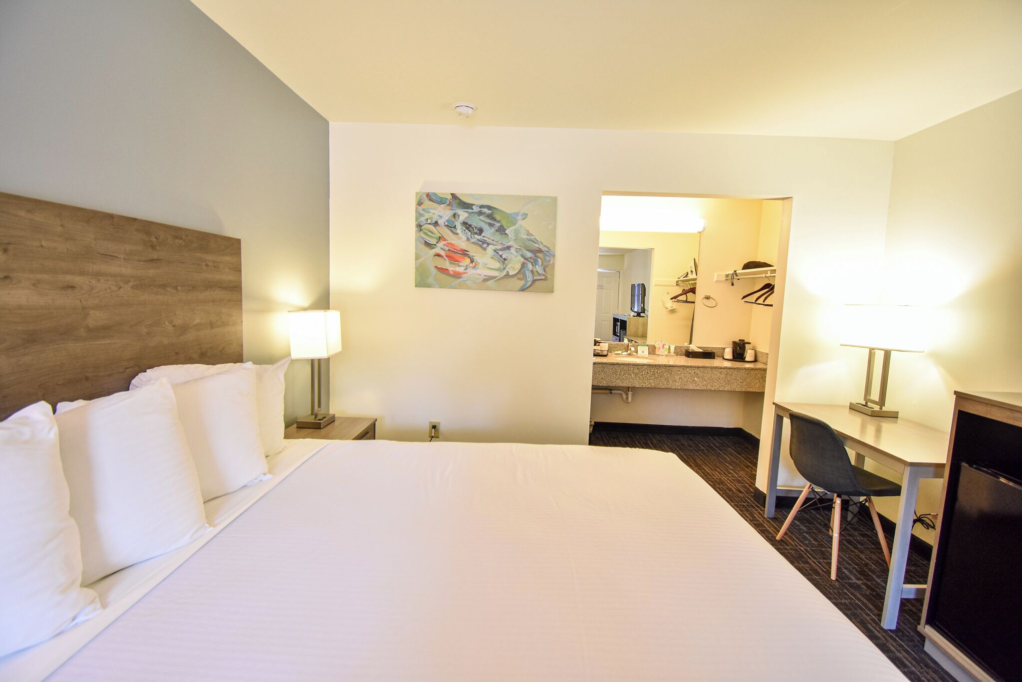 Una habitación de hotel con una cama grande, un escritorio con una silla, una lámpara de escritorio, un cuadro en la pared y un baño visible a través de una puerta abierta.