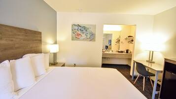 A hotel room with a large bed, a desk with a chair, a desk lamp, a painting on the wall, and a bathroom visible through an open door.