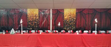 A banquet hall with a red tablecloth, white chairs, and a stage with red curtains.