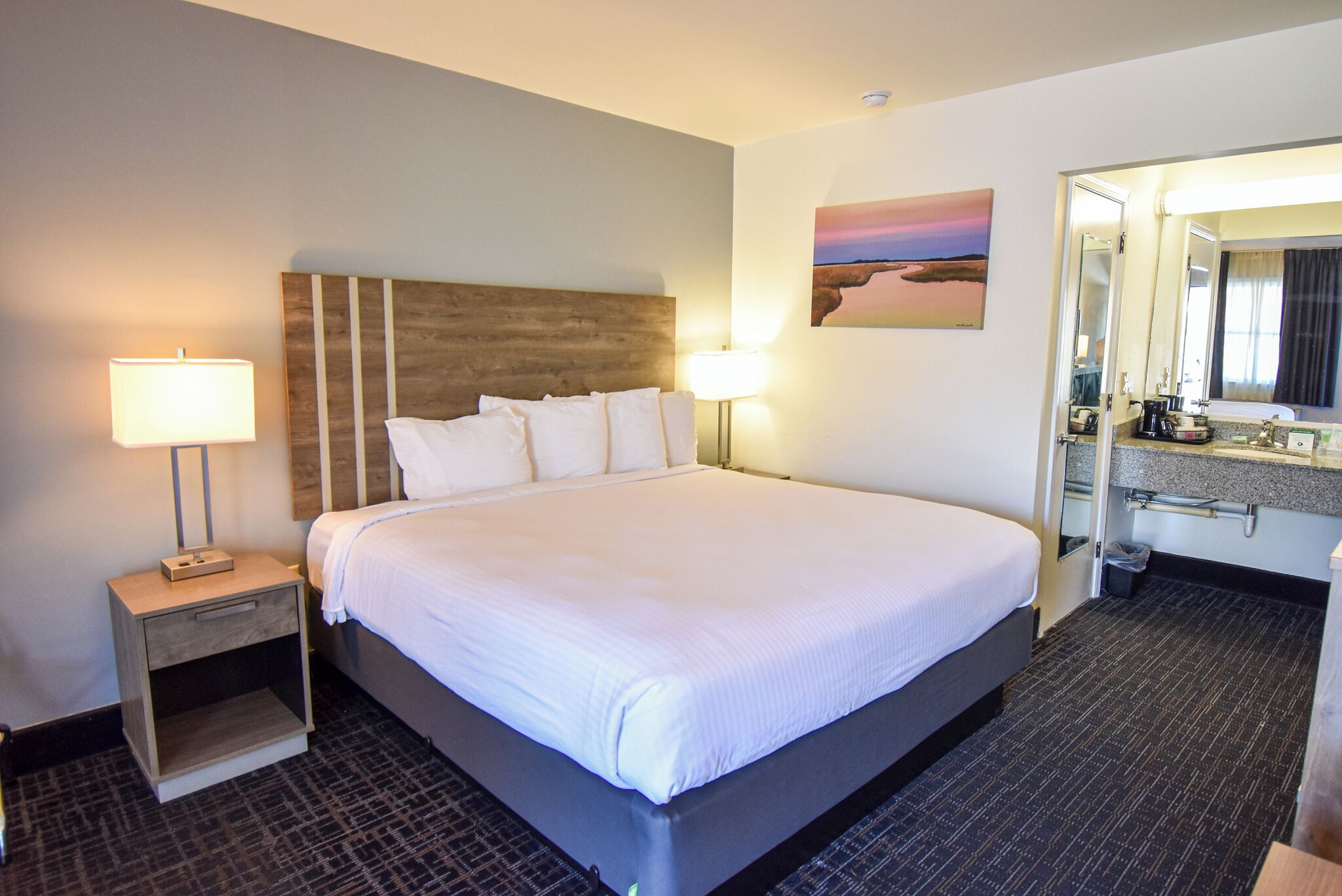 Hotelkamer met een groot bed, nachtlampjes, een nachtkastje, een badkamer met spiegel en een schilderij aan de muur.