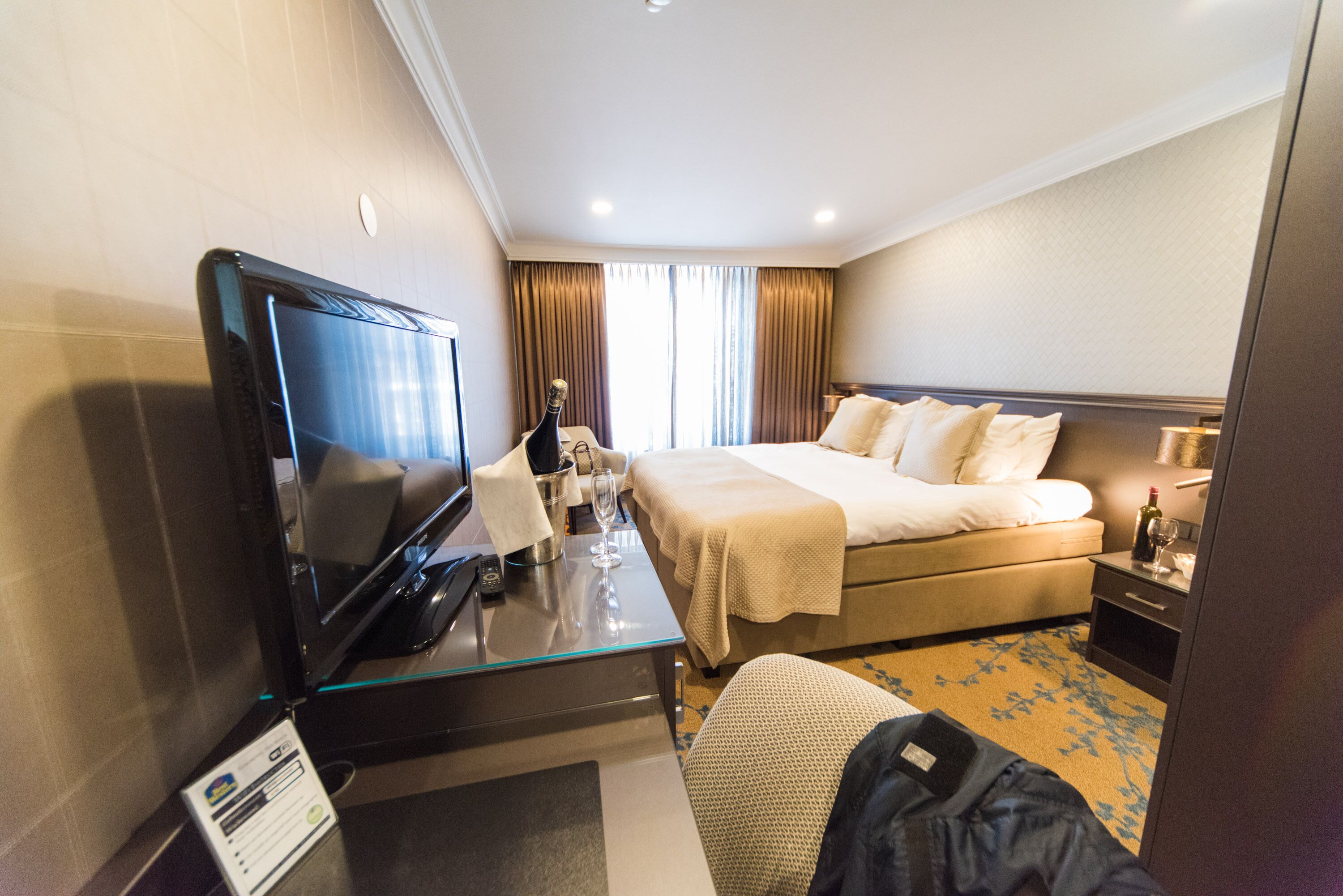 Una habitación de hotel con una cama, un televisor, un escritorio con una computadora, una botella de champán y una mesita de noche con una lámpara.