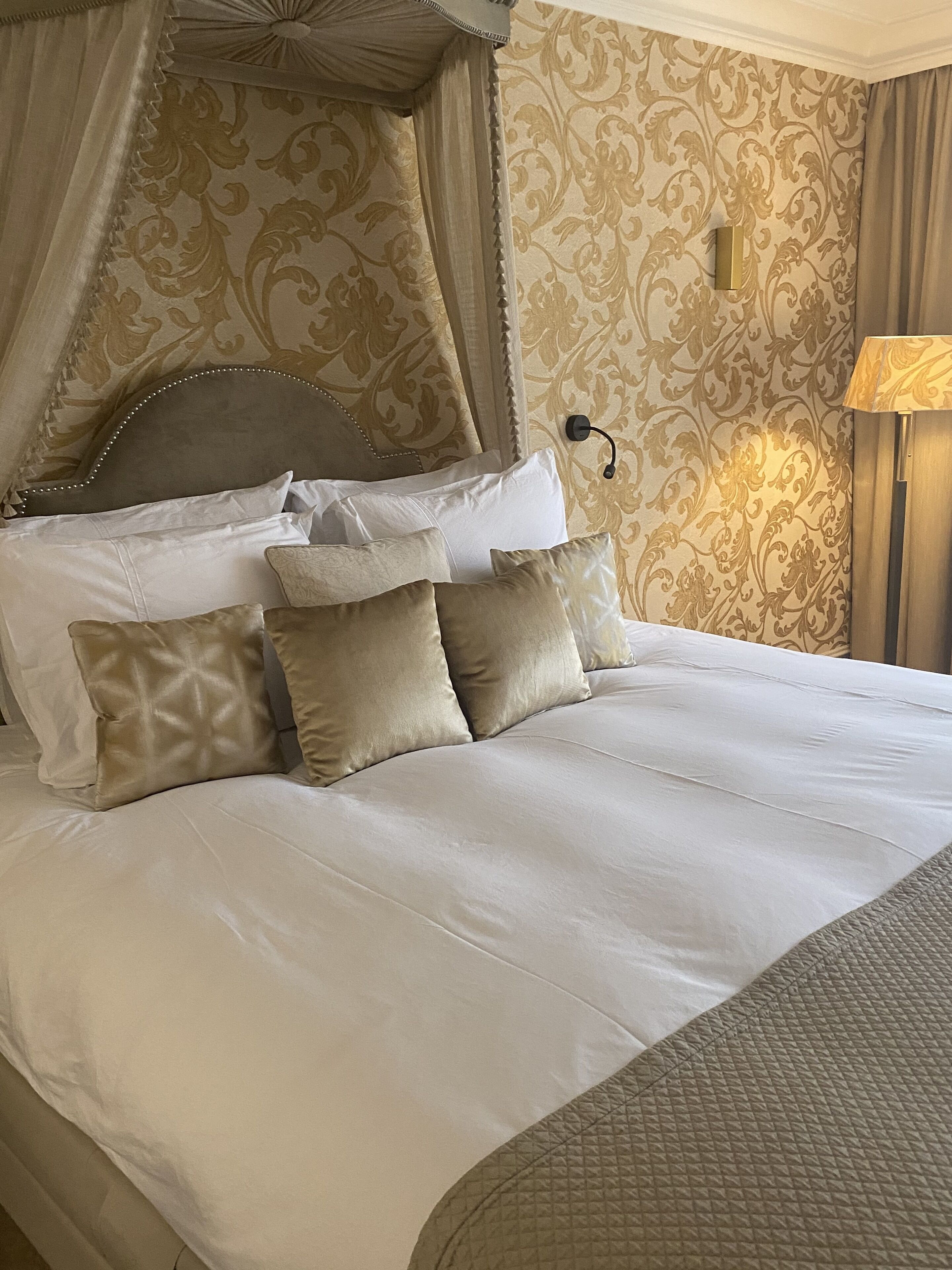 A four-poster canopy bed with multiple pillows, a headboard with bead trim, and a wall-mounted lamp.