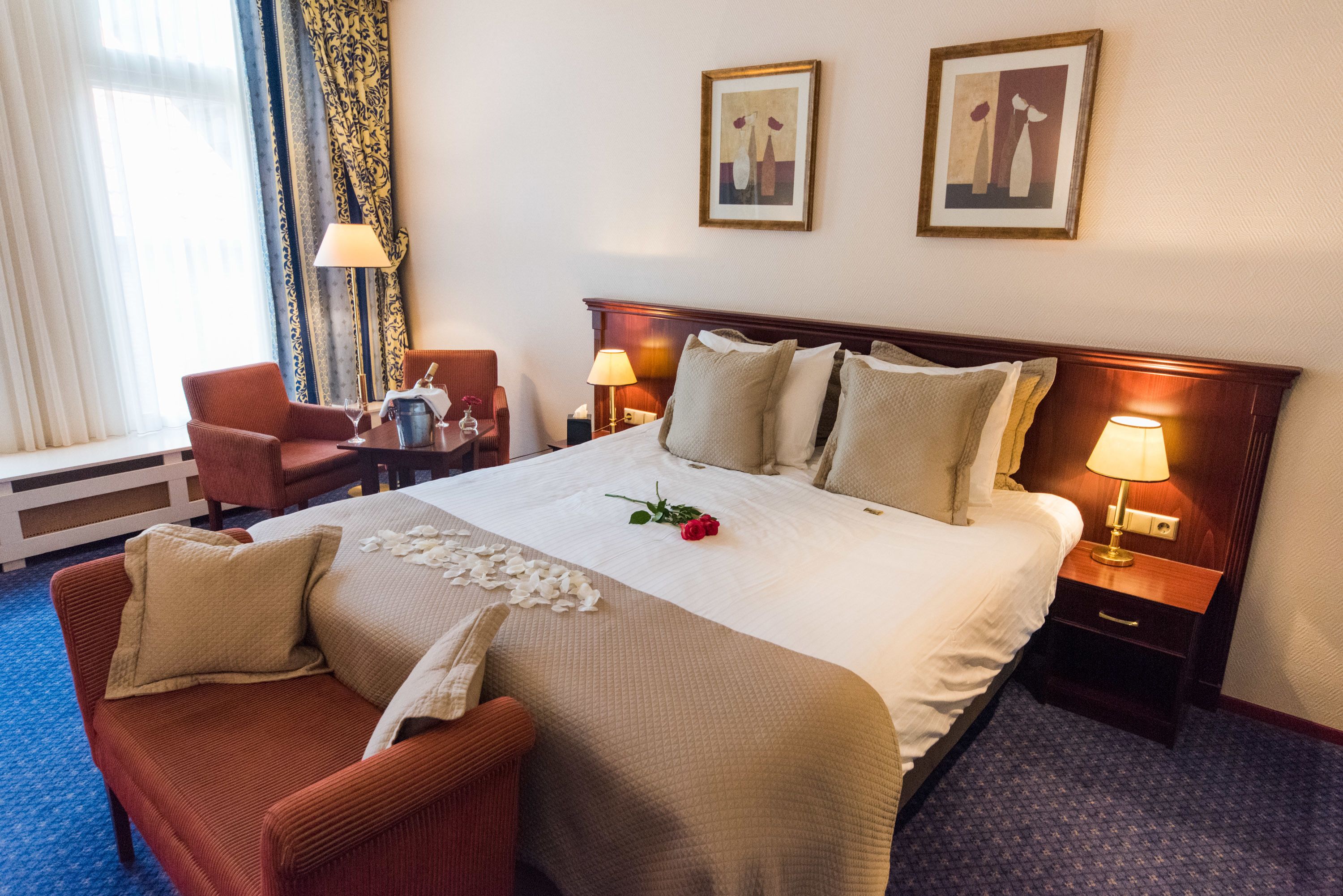 Een hotelkamer met een groot bed, twee nachtkastjes met lampen, een stoel, een bureau met lamp en een raam met gordijnen.