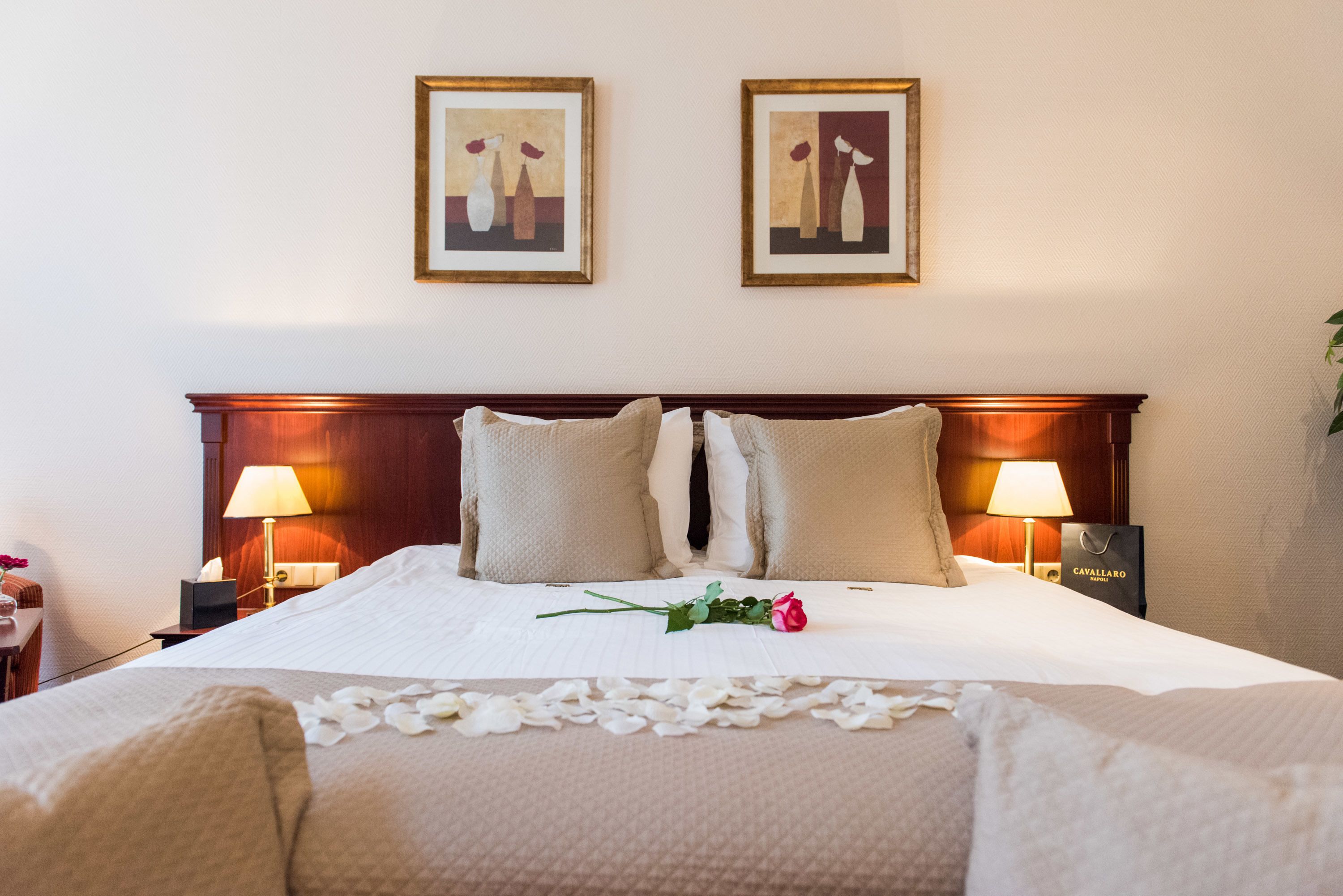 Een hotelkamer met een groot bed, twee bedlampen, een lijst met kunstwerk erboven en een decoratieve roos op het beddegoed.