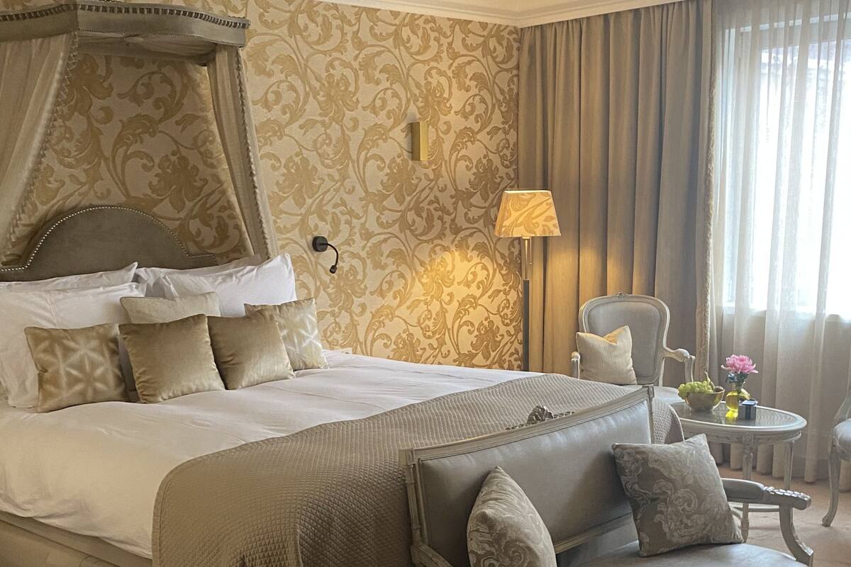 Une chambre spacieuse avec un grand lit, une chaise longue, une petite table ornée de fleurs et une lampe.