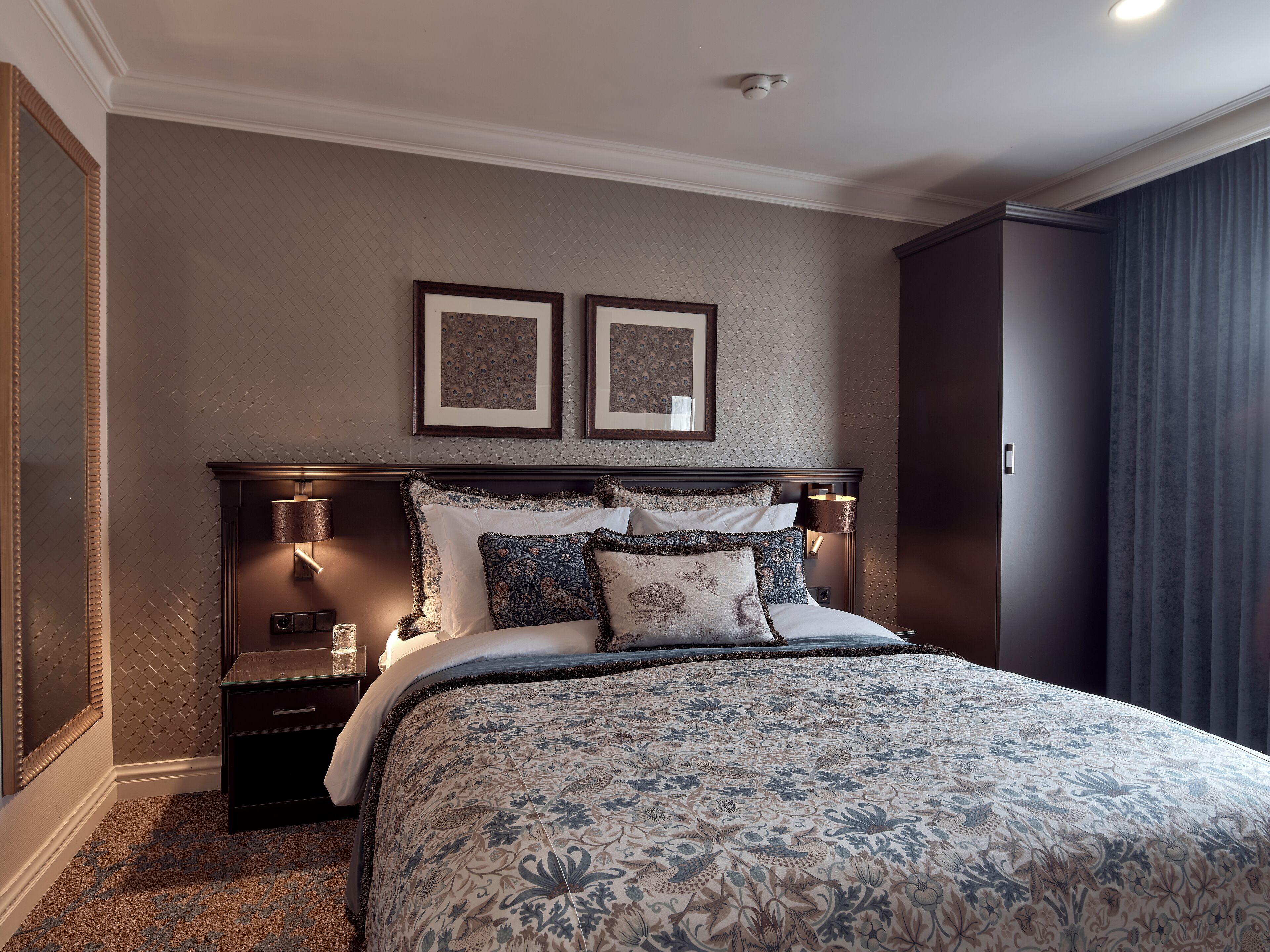 Een net opgemaakt bed met een bloemenprint deken, twee ingelijste foto’s aan de muur erboven en een donker hoofdboard.