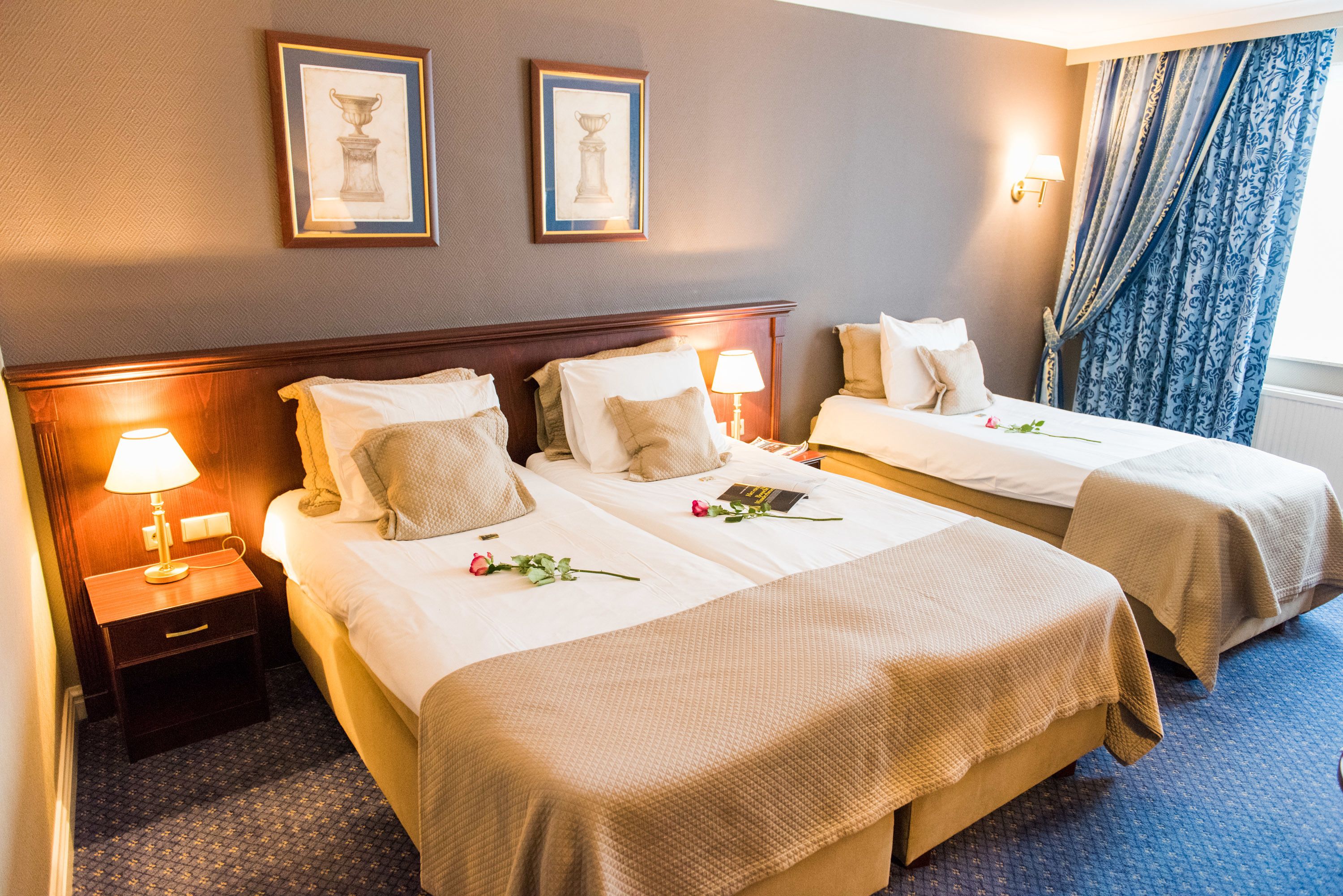 A hotel room with two beds, a nightstand with a lamp, and two framed pictures on the wall.