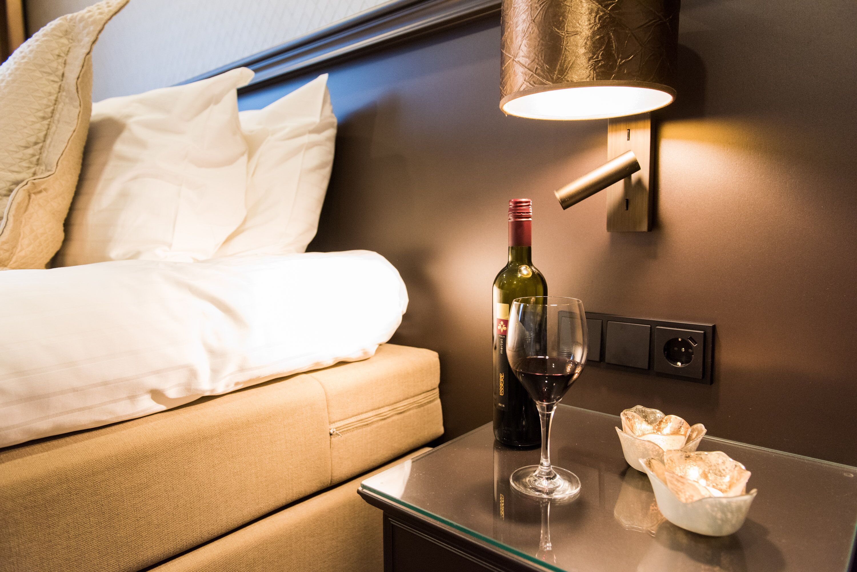 A neatly made bed with white linens, a bottle of wine, a wine glass, and two small bowls of bread on a bedside table.
