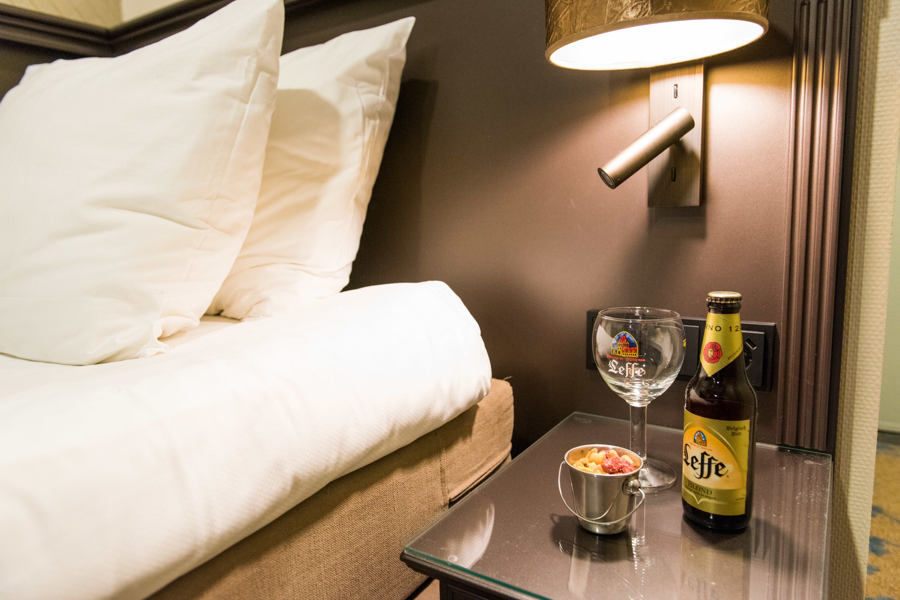 A hotel room with a bed, a bedside table with a bottle of Leffe beer, a glass, and a small bowl of snacks.