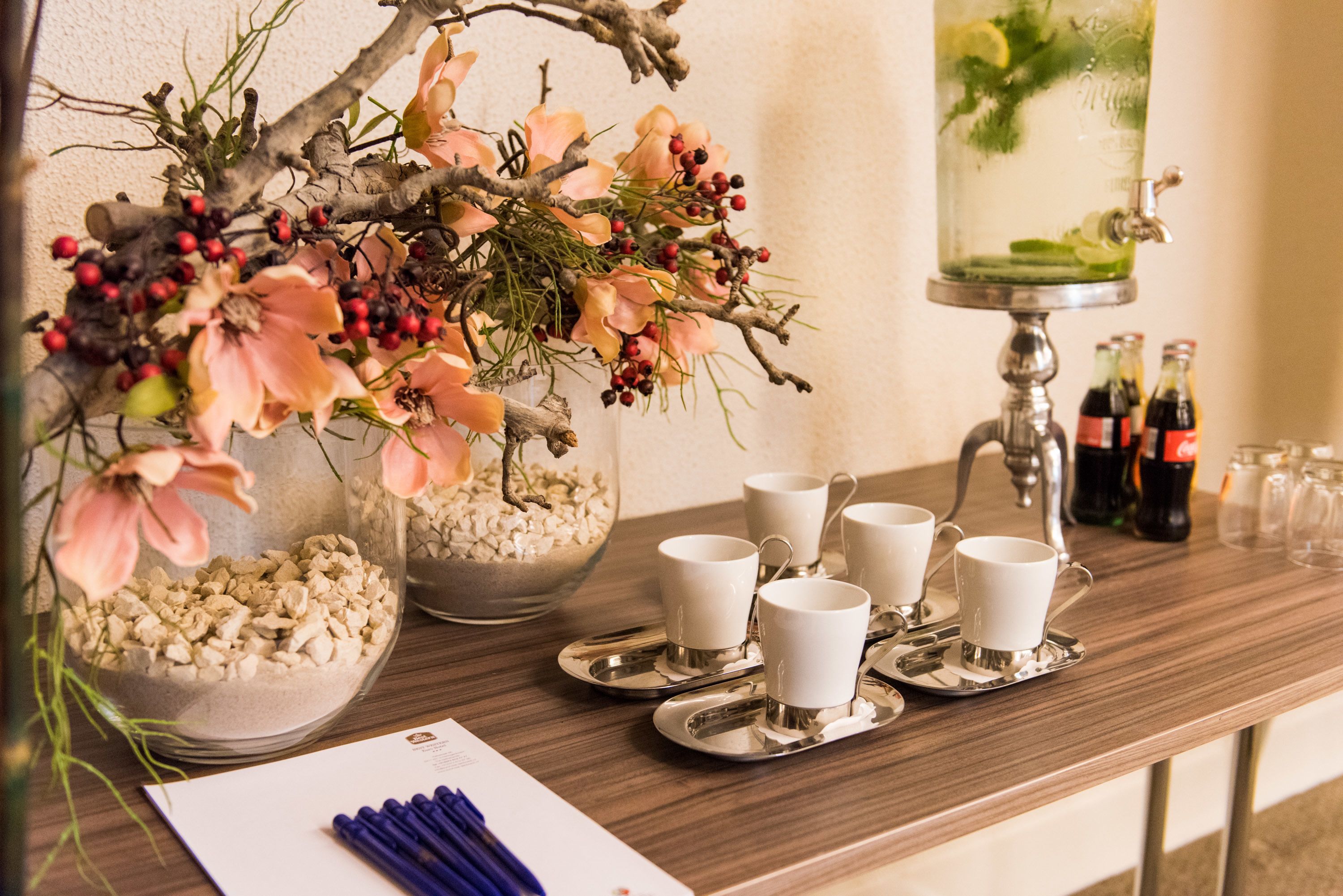 A wooden table with a floral arrangement, a glass container with a drink, and several white cups and saucers.