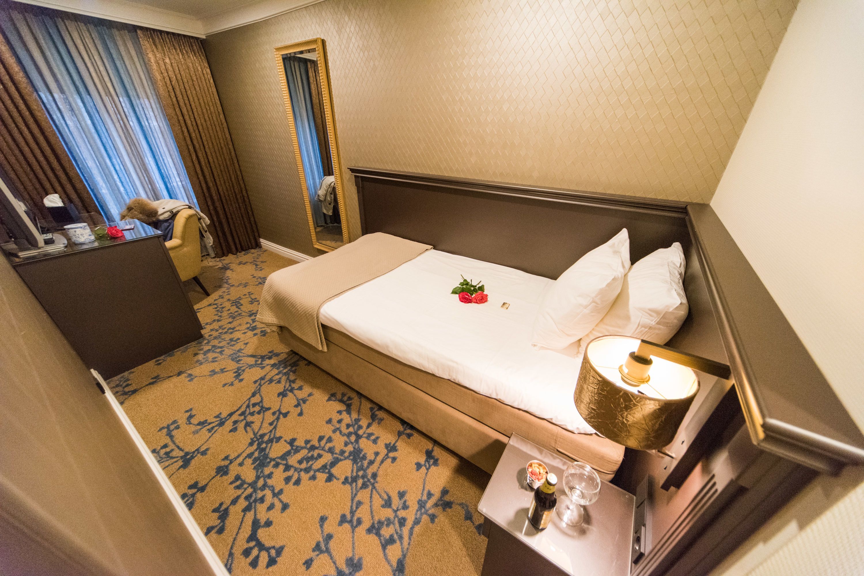 A hotel room with a bed, a desk, a mirror, and a nightstand with a lamp.
