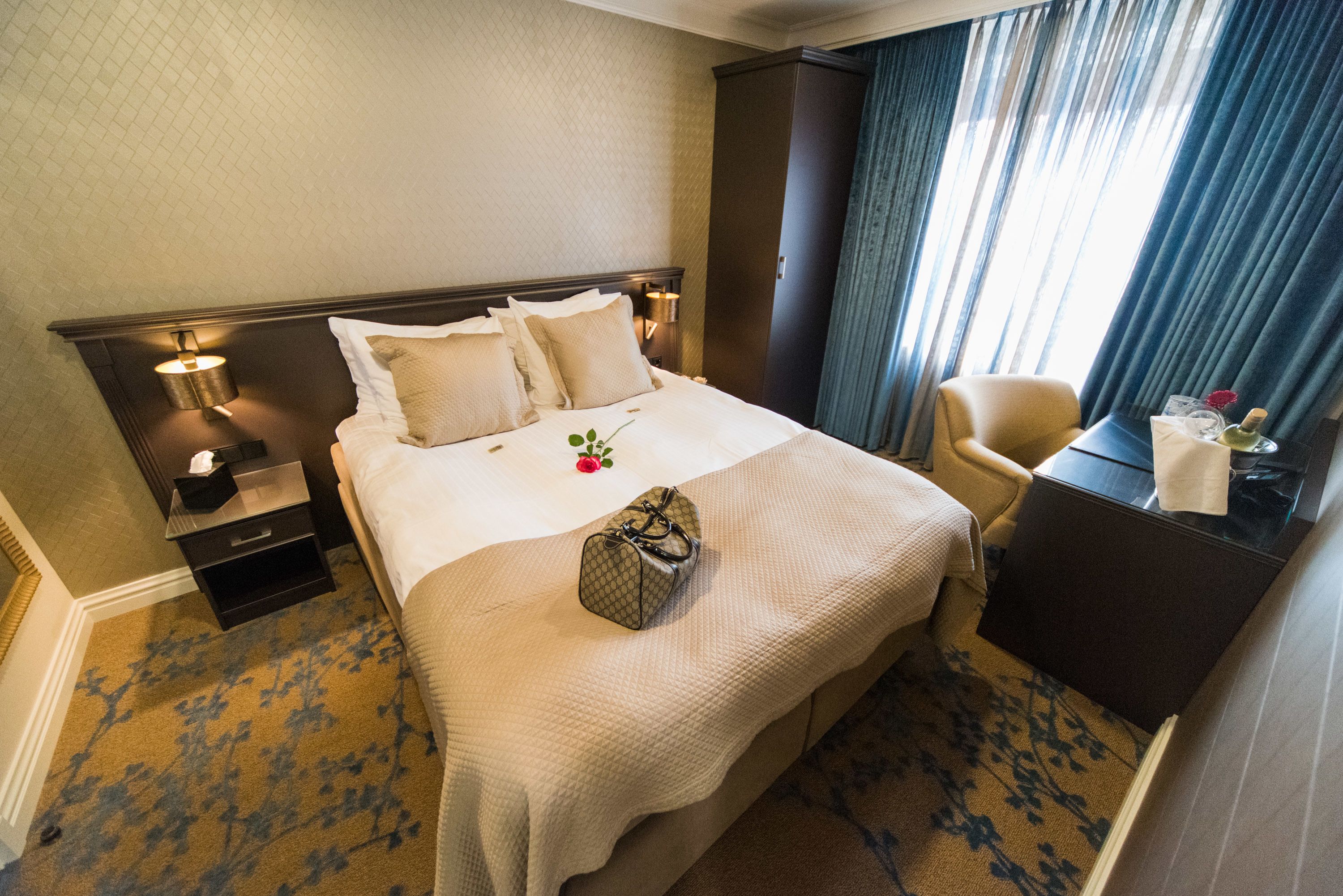 A hotel room with a large bed, a nightstand, a chair, a desk, and a window with curtains.
