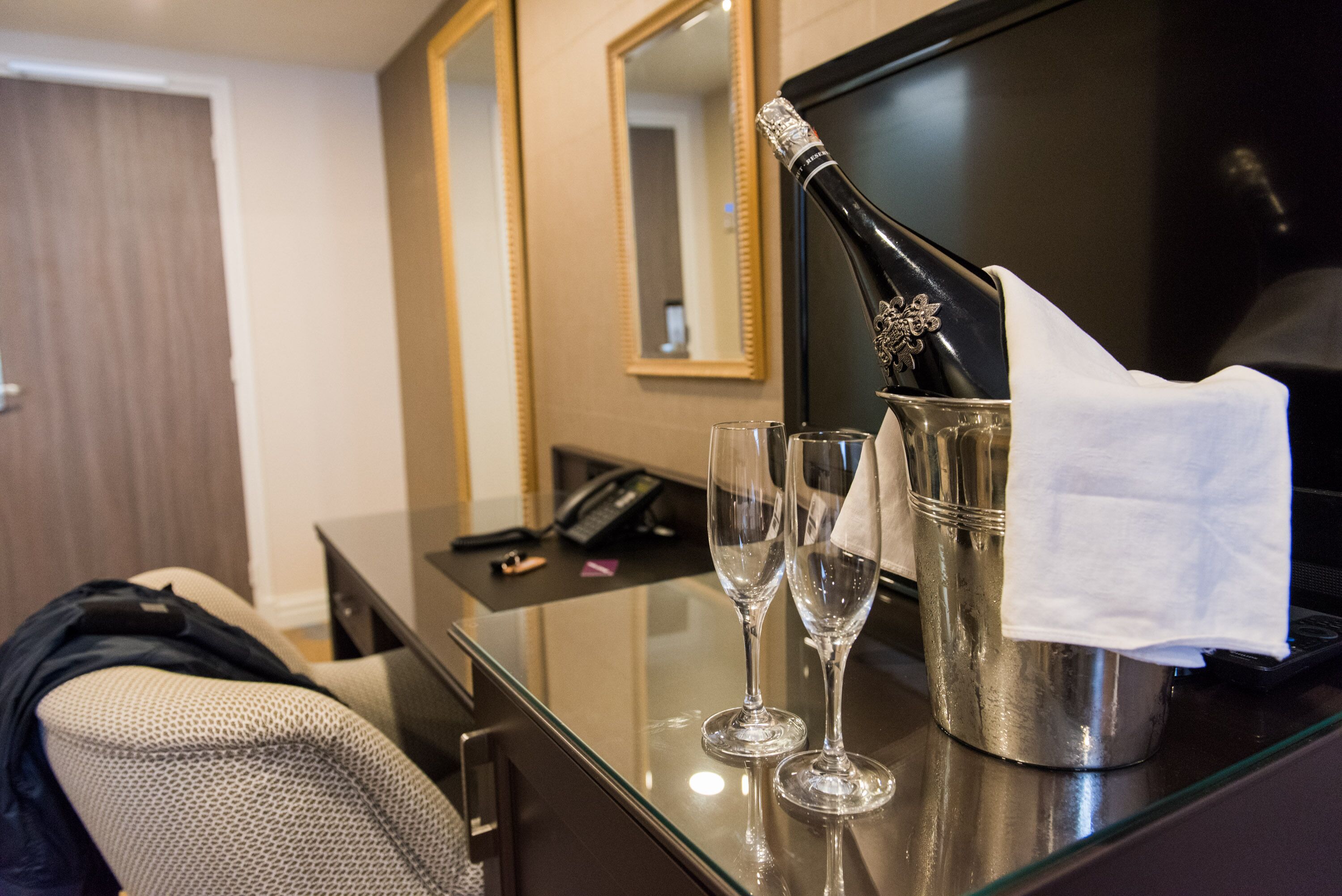 A hotel room with a minibar, a mirror, a desk with a phone, and two empty wine glasses.