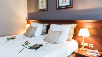 A neatly made bed with white linens, two pillows, a bedside lamp, a magazine, and a rose on the bed.