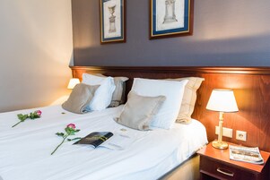 A neatly made bed with white linens, two pillows, a bedside lamp, a magazine, and a rose on the bed.