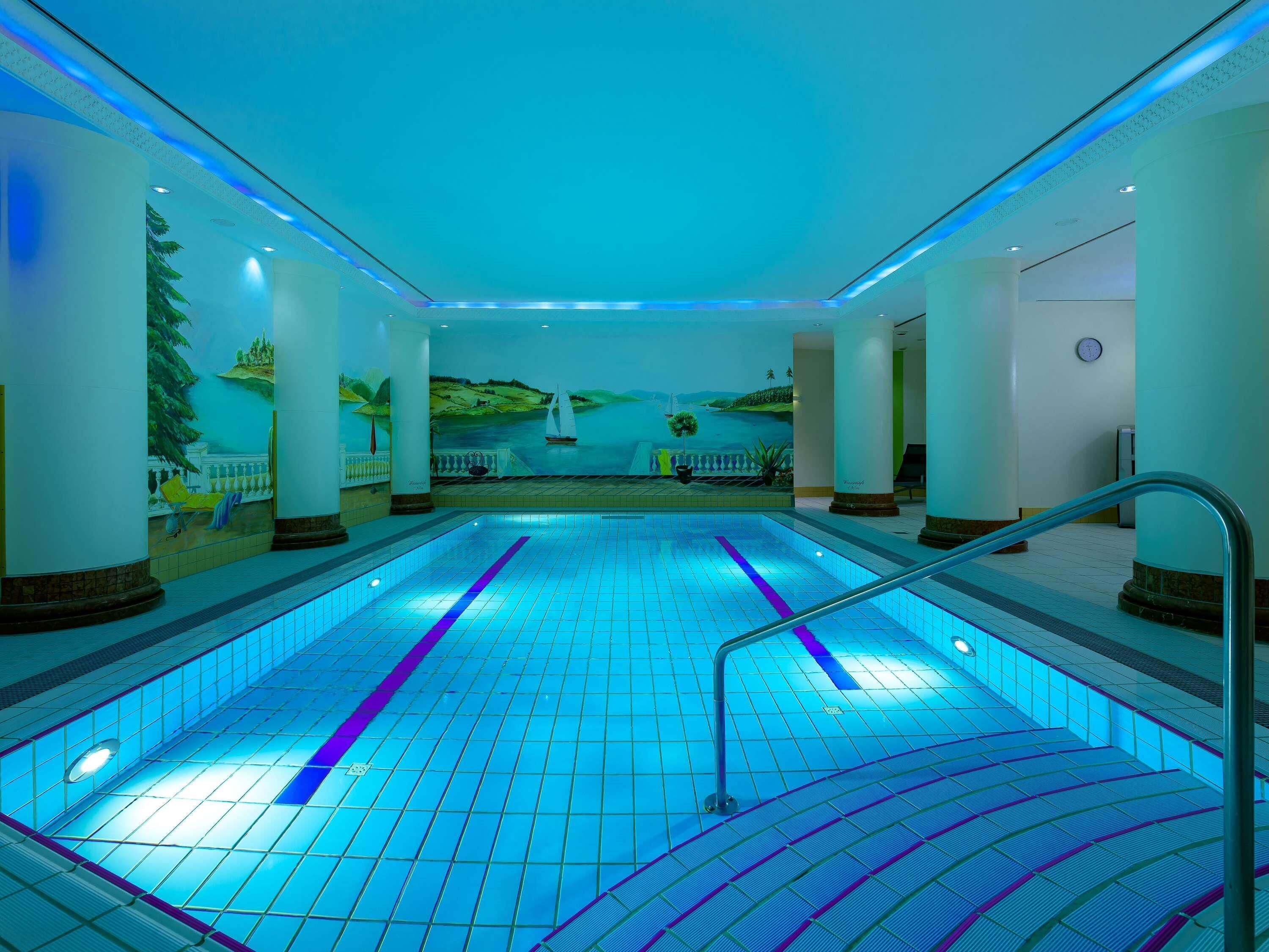 Ein Schwimmbecken mit Fliesenboden, einer Wandmalerei, die eine Landschaft zeigt, und einer Uhr an der Wand.