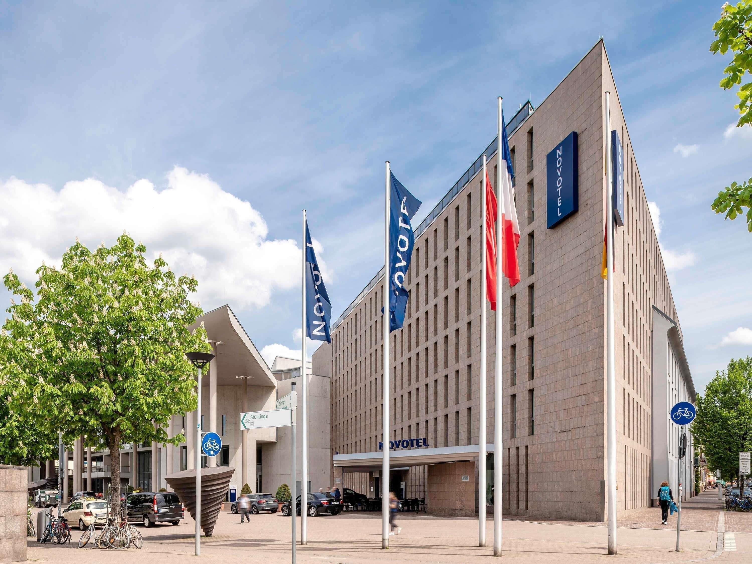 Un hôtel moderne, reconnaissable à son grand panneau « NOVOTEL », à ses nombreux drapeaux et à l’espace clairement délimité pour le stationnement des vélos.