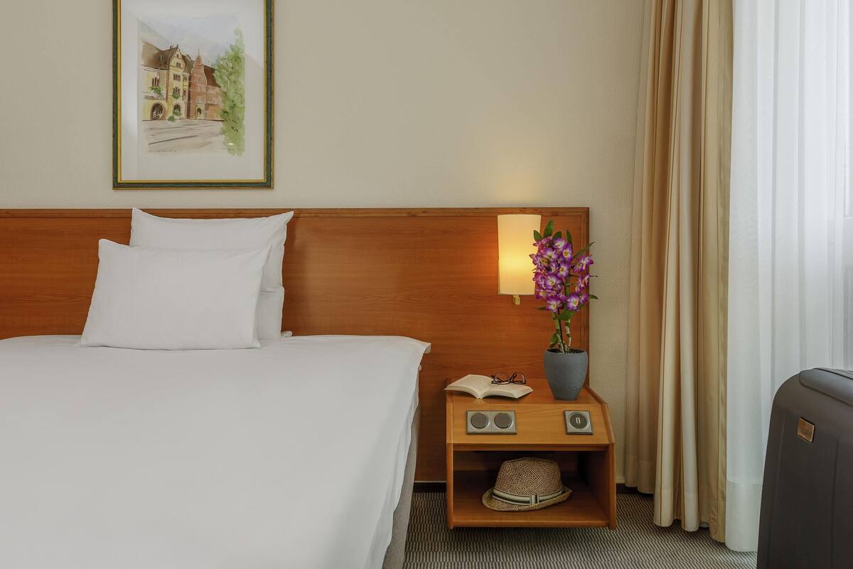 Een hotelkamer met een bed, een nachtkastje met een lamp, een vaas met bloemen, een hoed en een ingelijst foto op de muur.