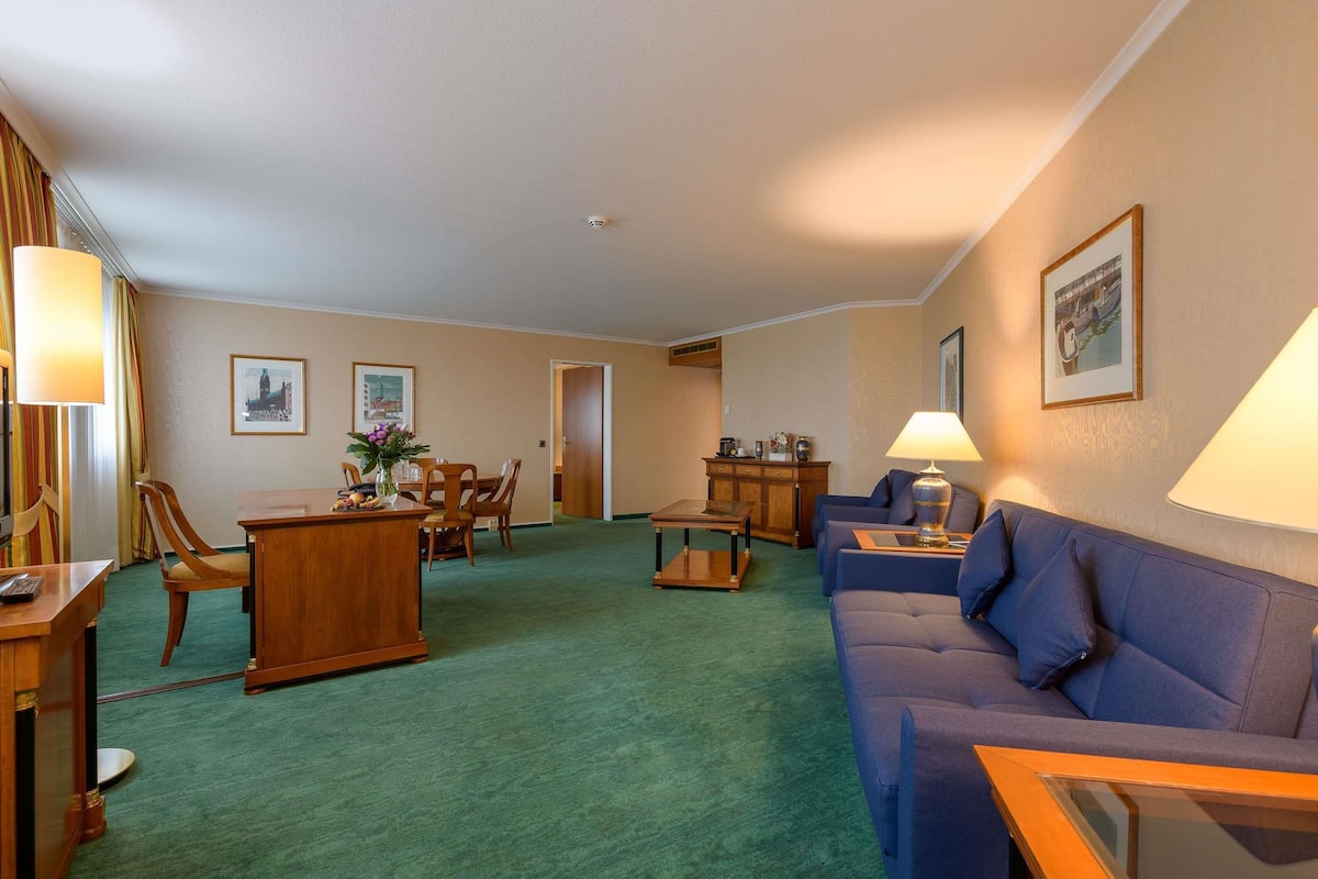 Een hotelkamer met een groen tapijt, een houten bureau, een blauwe bank en een zithoek met stoelen en een tafel.