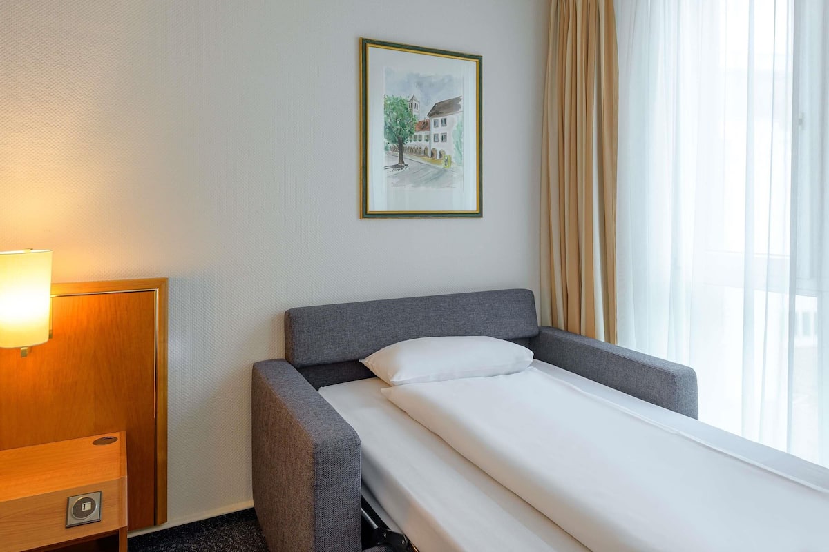 Een hotelkamer met een bed, nachtkastje, lamp, raam met gordijnen en een lijst aan de muur.