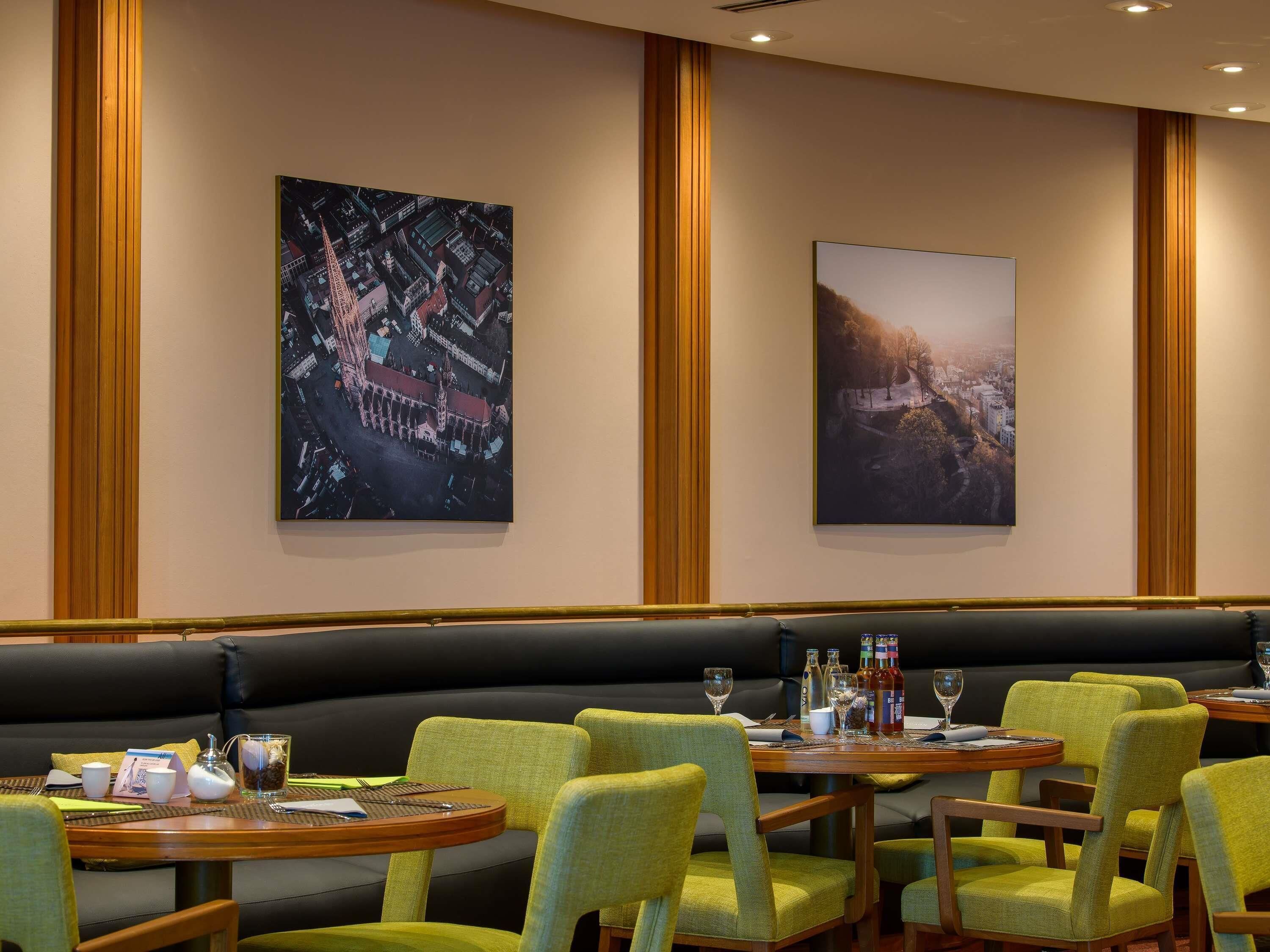 A restaurant with green upholstered booths, round tables, and two large framed cityscape paintings on the wall.