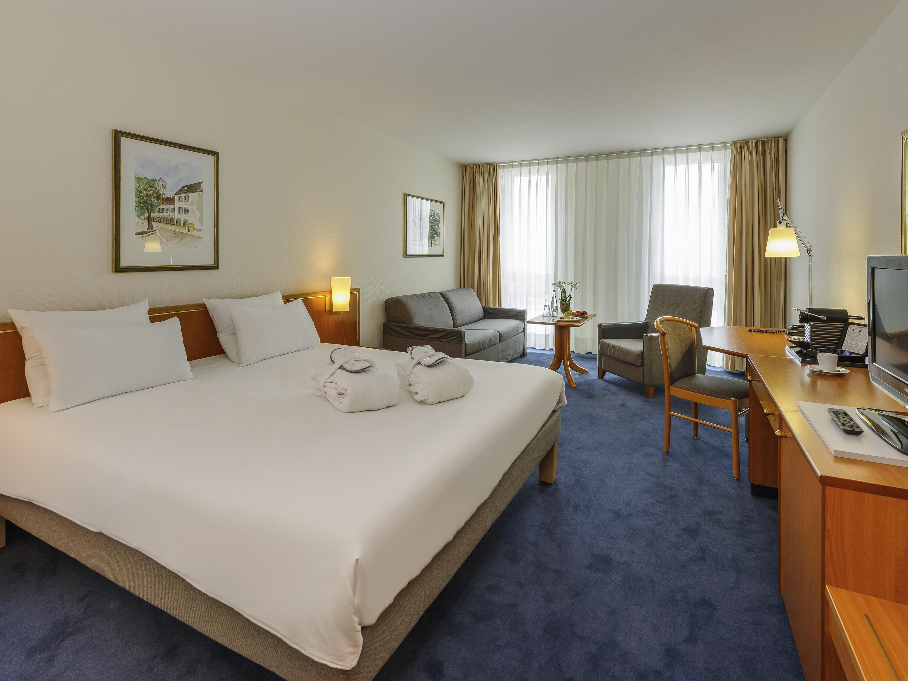 Une chambre d’hôtel avec un grand lit, deux fauteuils, un bureau avec une télévision et une fenêtre avec des rideaux.